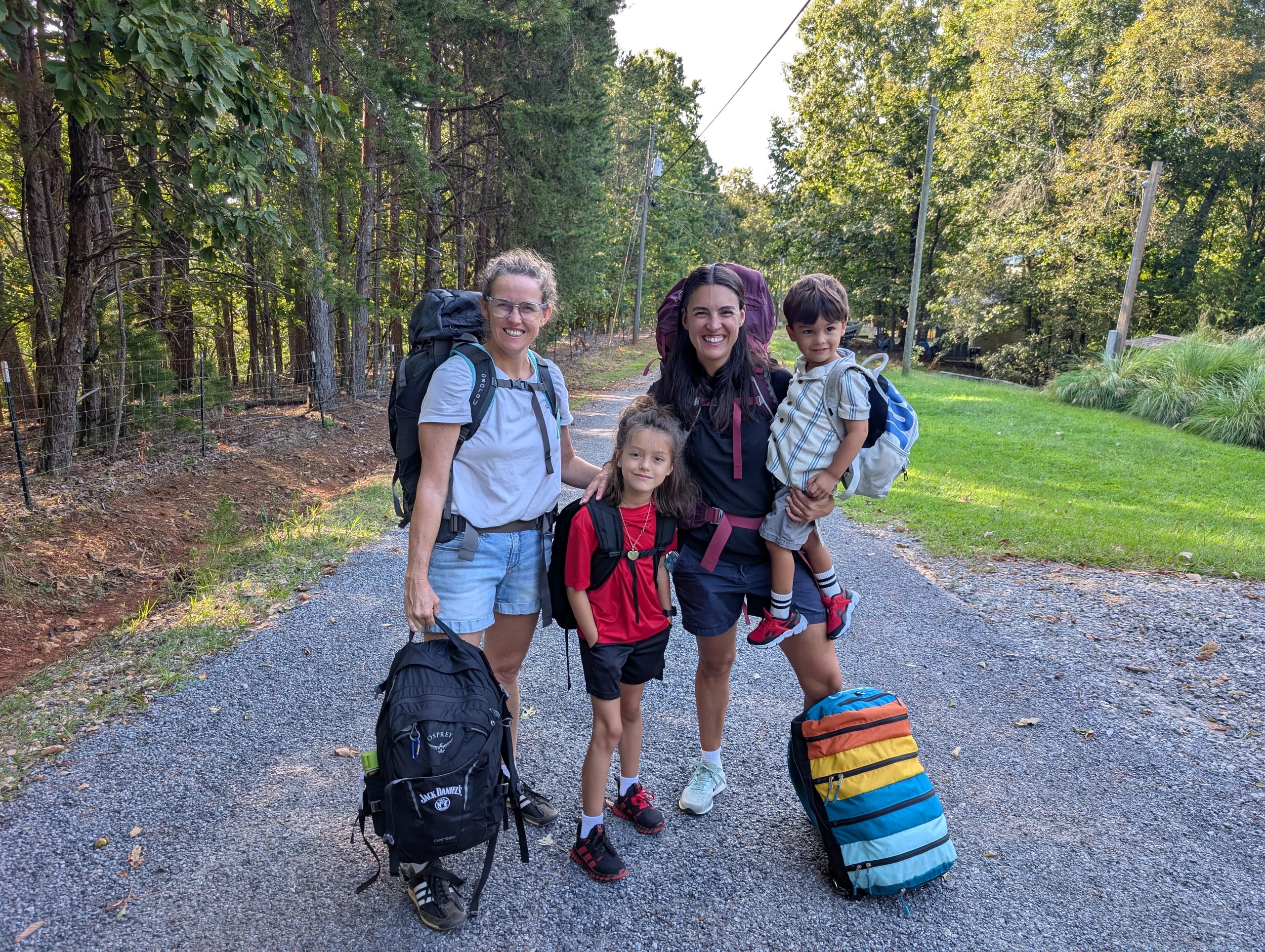 Alissa, Dani, Remi and Arlo standing on a gravel driveway with backpacks on, ready to leave for the trip