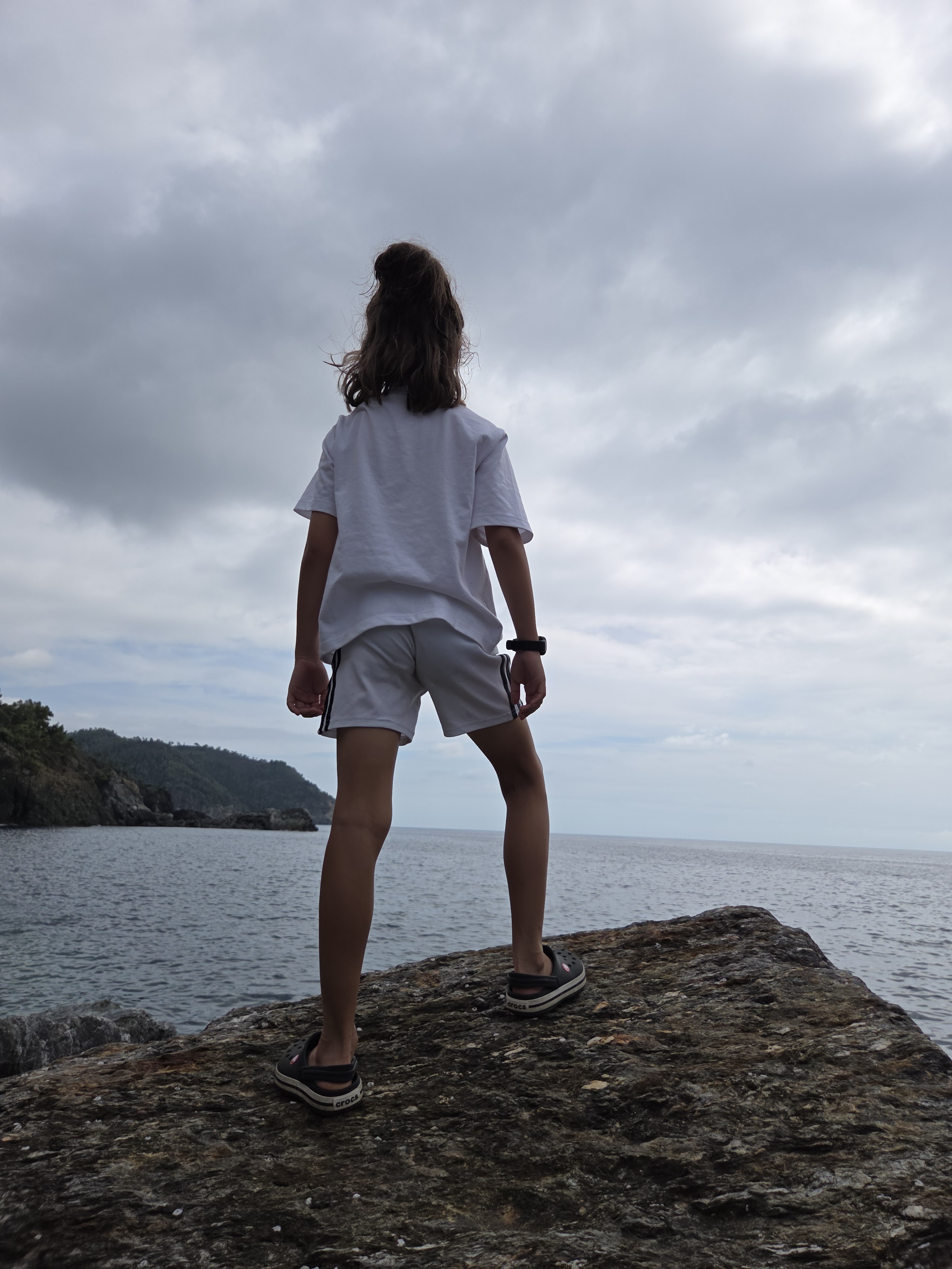 Remi standing on coastal rocks from behind, looking out over the Ligurian sea toward a wooded headland under a cloudy sky