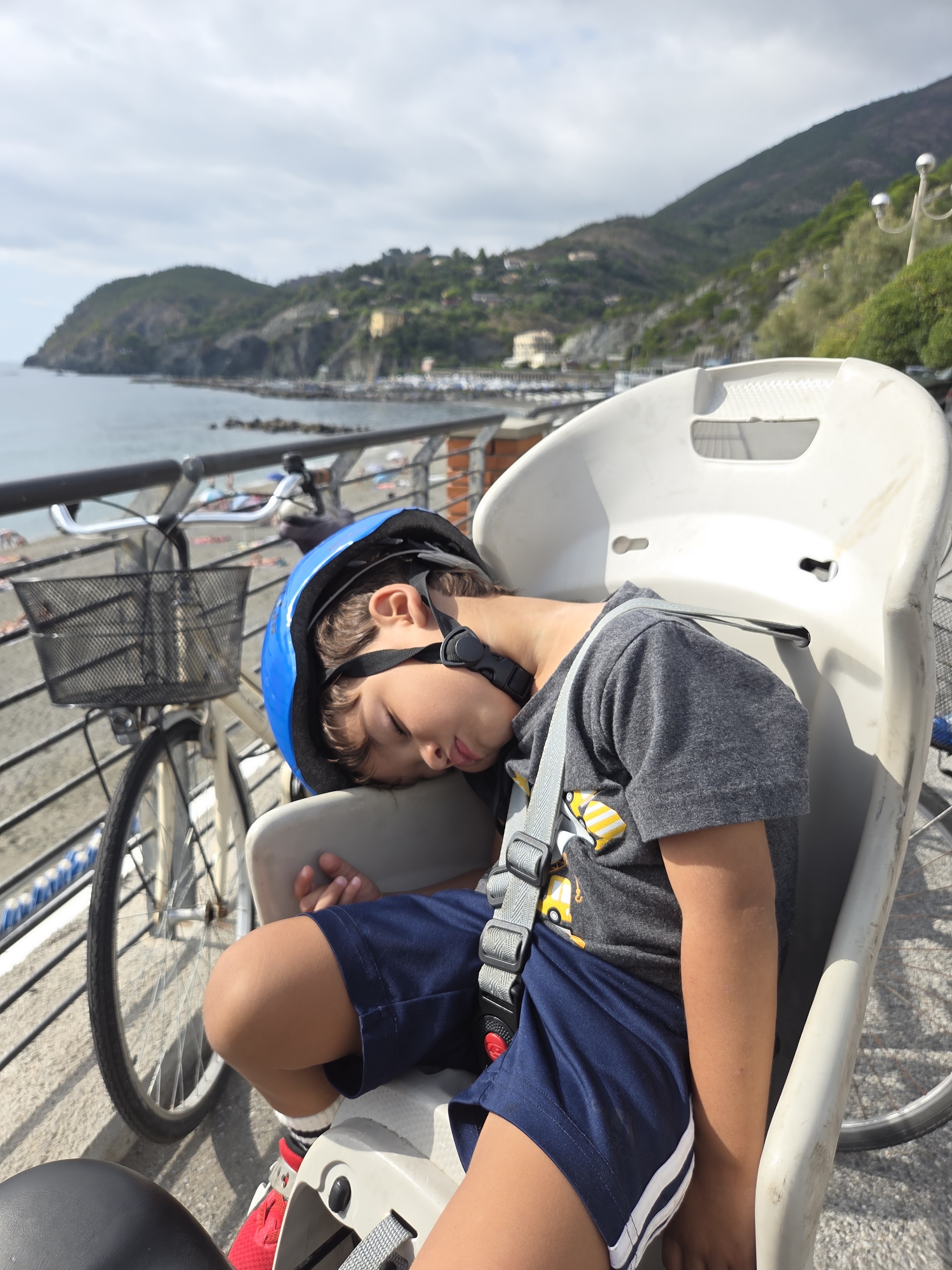 Arlo asleep in the rear bike seat with his helmet on, head resting on the seat back, Levanto's beach and green hillside visible behind him