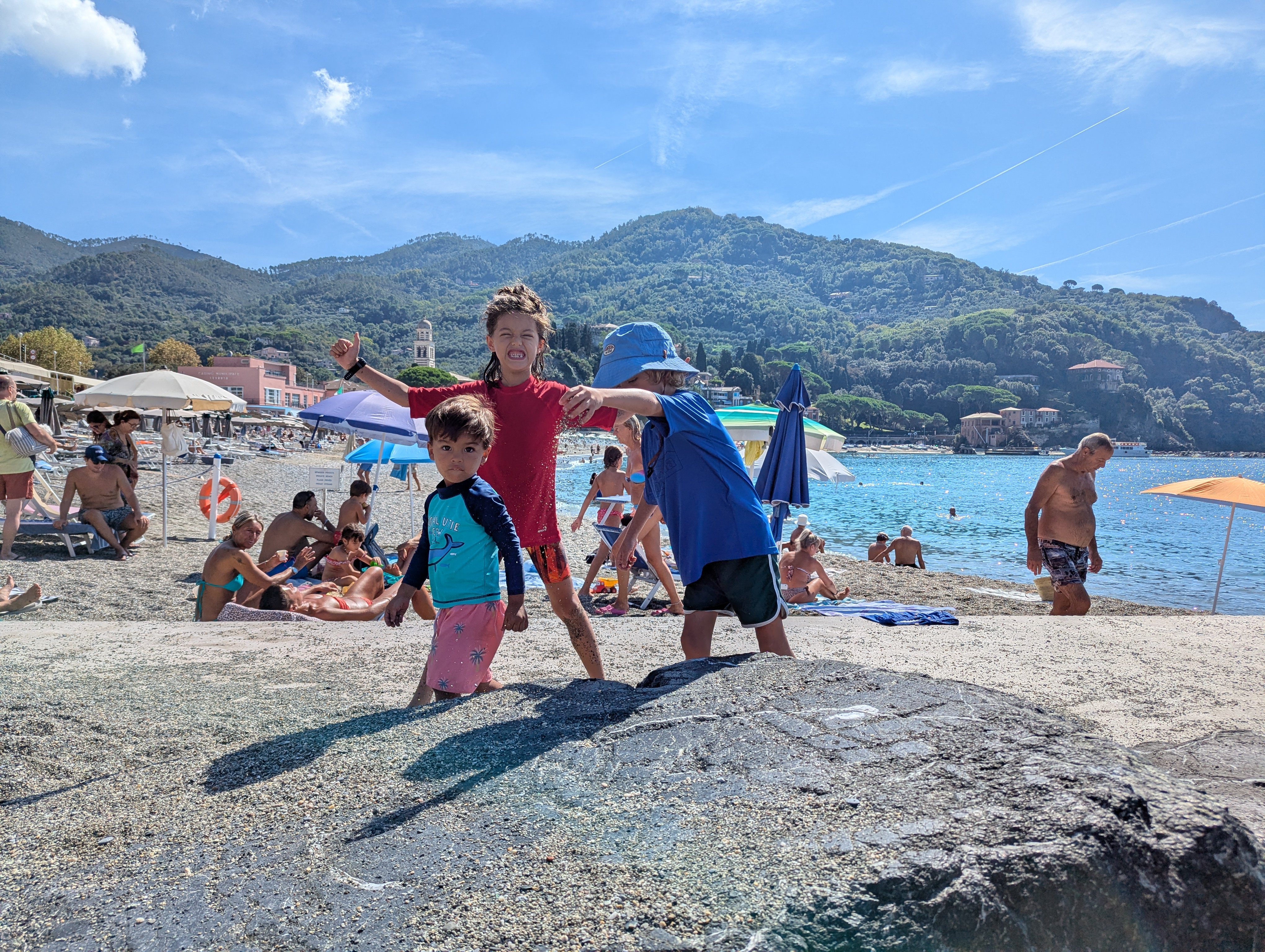 Remi, Arlo and River standing together on a large flat rock at Levanto's beach, Remi grinning with arms spread wide, umbrellas and mountains behind them