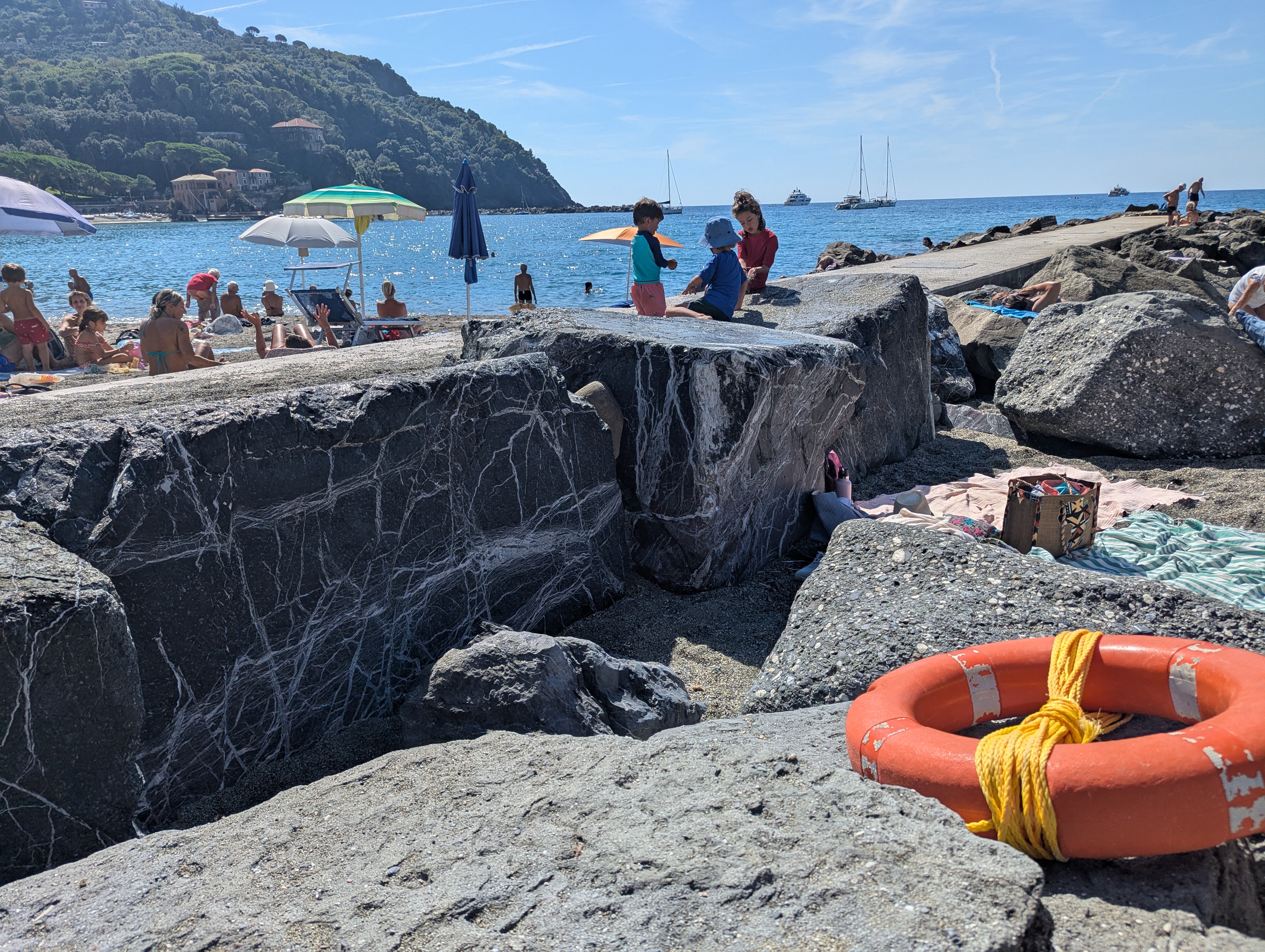Remi, Arlo and River crouched together on the pier's rock breakwater, examining something between the rocks, sailboats visible in the water behind them