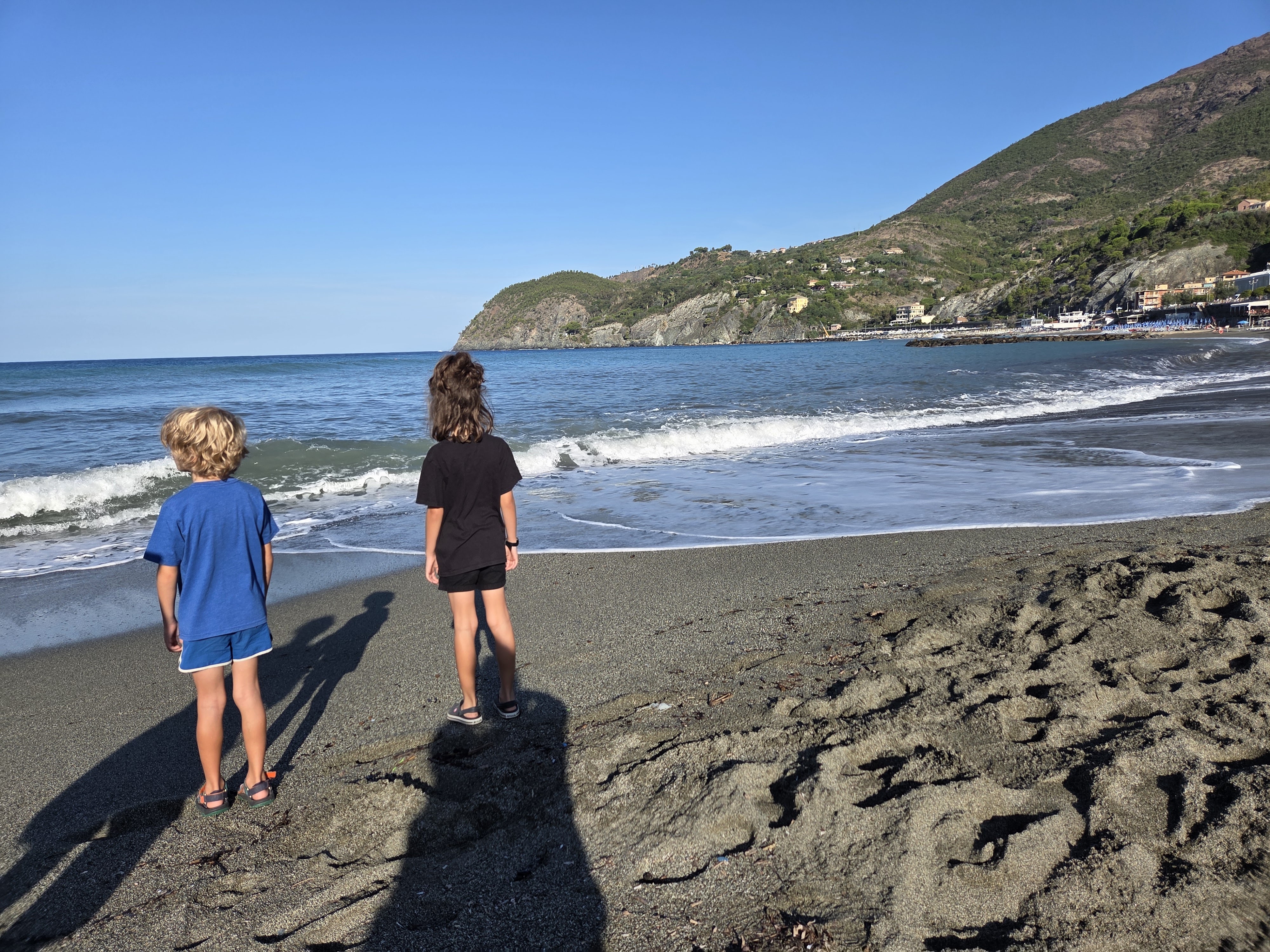 River and Remi standing at the edge of the water on Levanto's beach in the morning light, a headland visible across the bay
