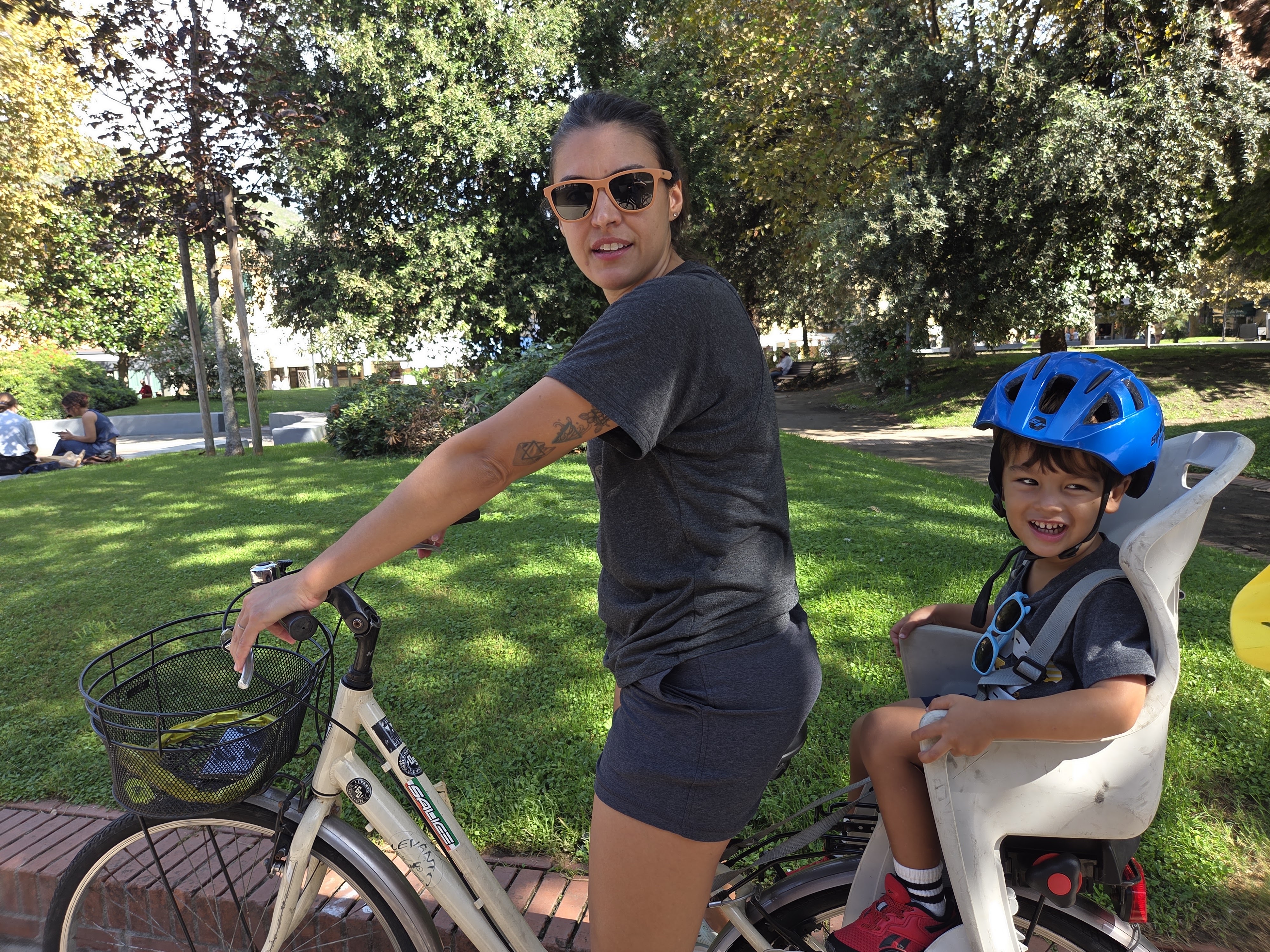 Alissa with Arlo in a rear bike seat, both smiling in a sunny park at the start of the bike trail