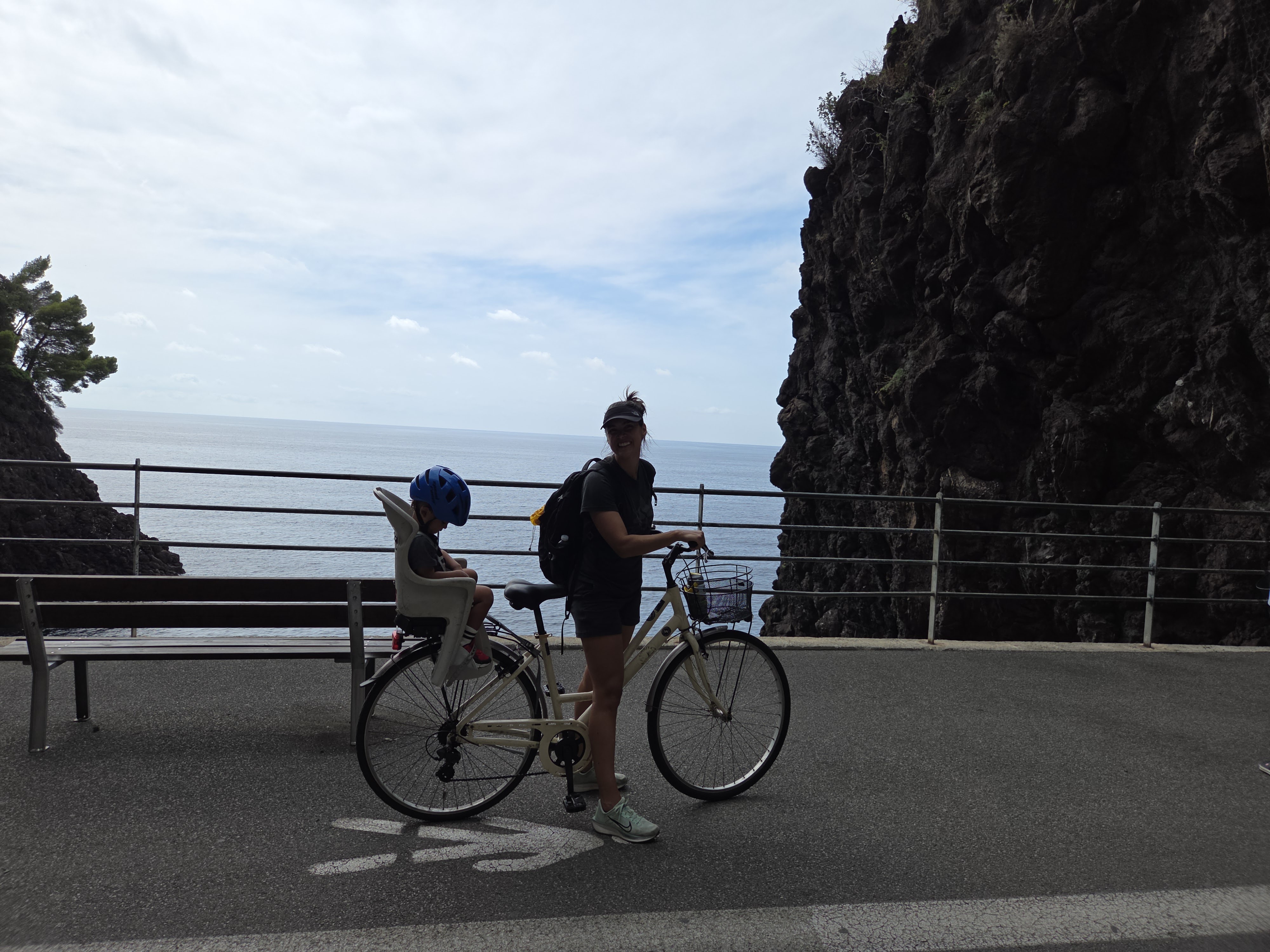 Alissa standing with Arlo in the rear bike seat on the coastal path, rugged black cliff on one side and open Mediterranean sea behind them