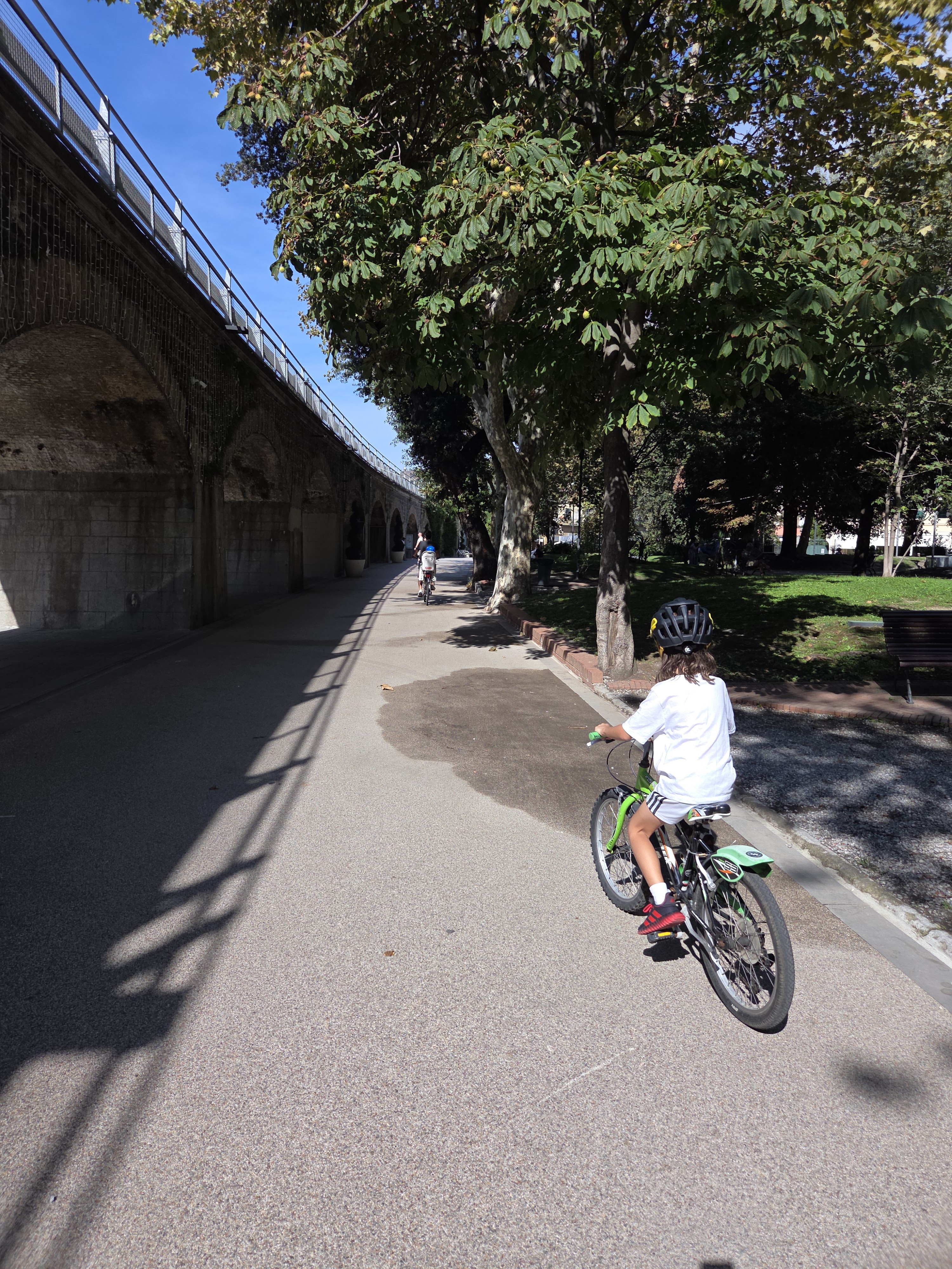 Remi biking along a wide path with a stone railway viaduct running alongside, trees and park on the right