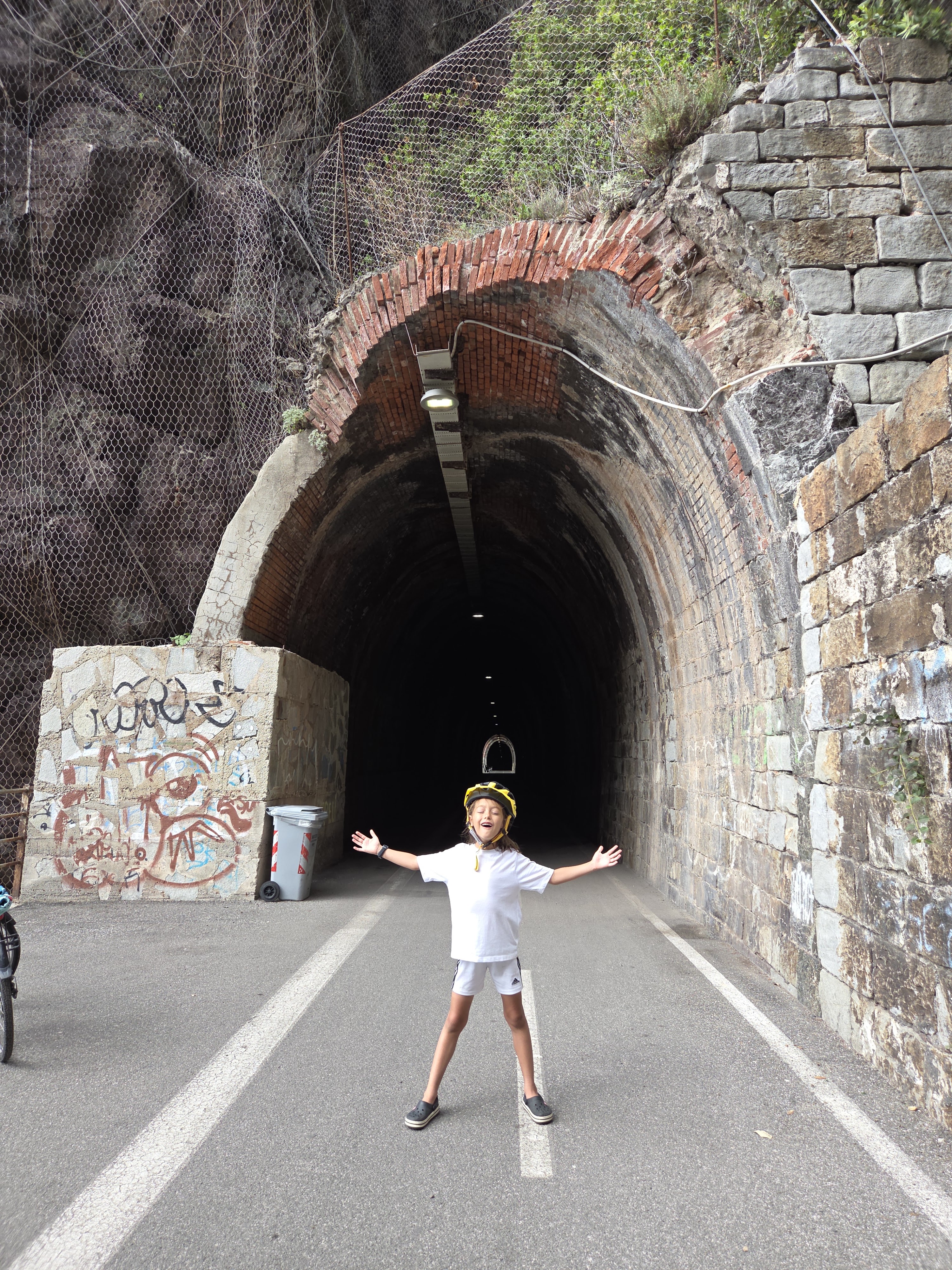 Remi standing with arms spread wide at the entrance of a brick-arched railway tunnel, lights stretching into the darkness