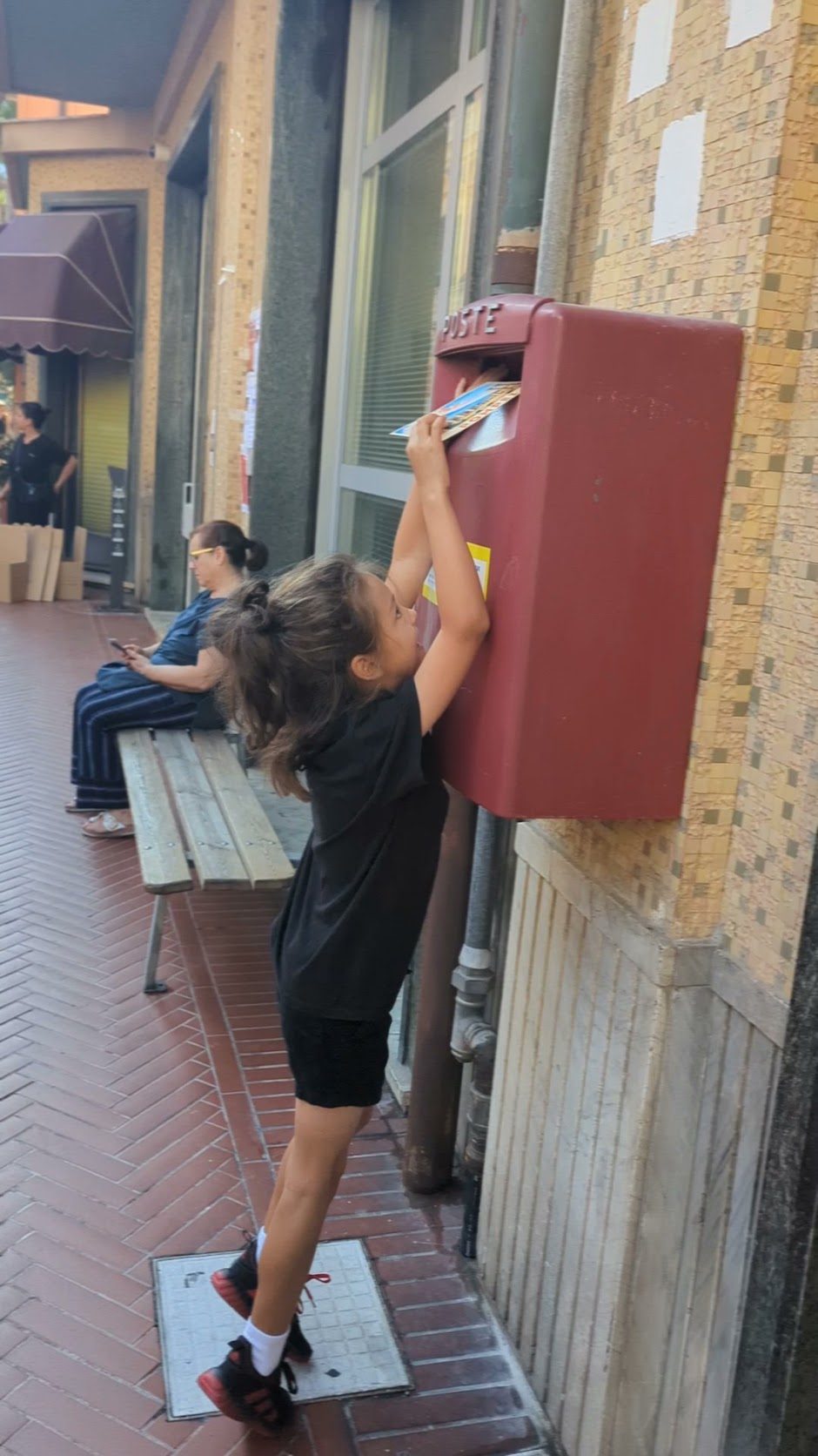 Remi reaching up on her toes to drop a postcard into a red La Poste mailbox on a street in Levanto