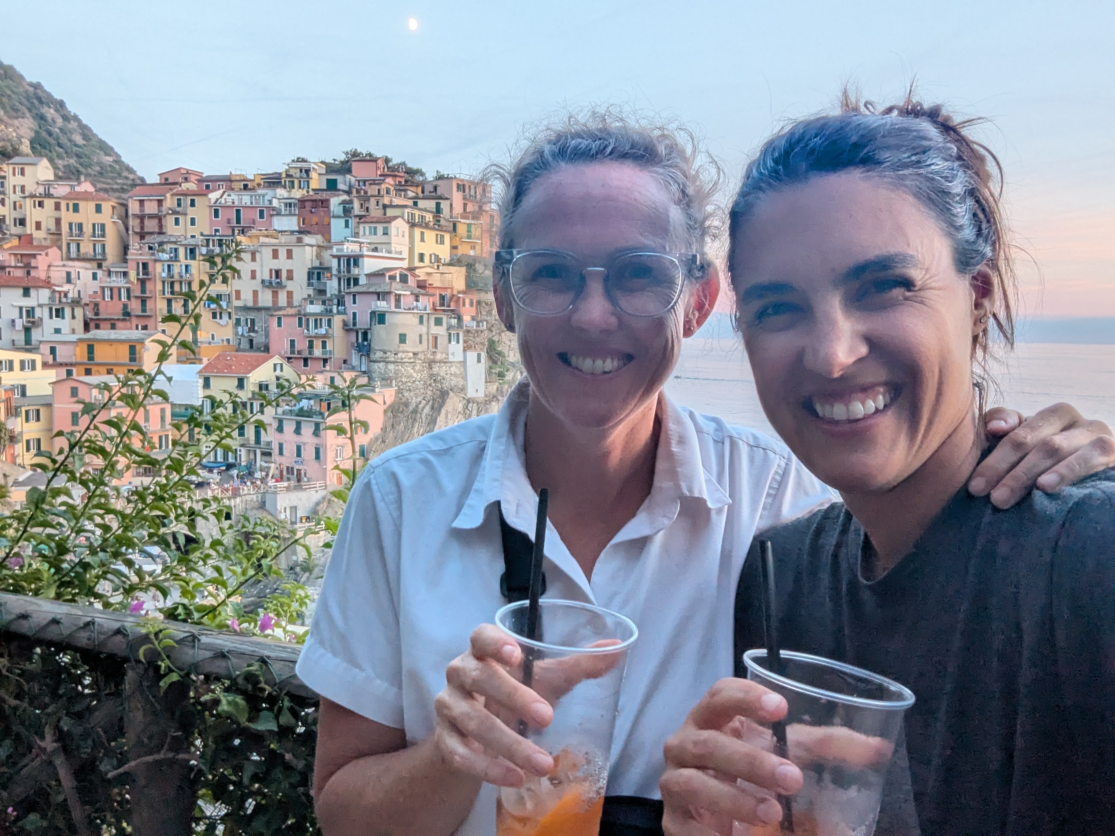 Alissa and Dani smiling with aperol spritzes in hand, Manarola's colorful cliffside village behind them