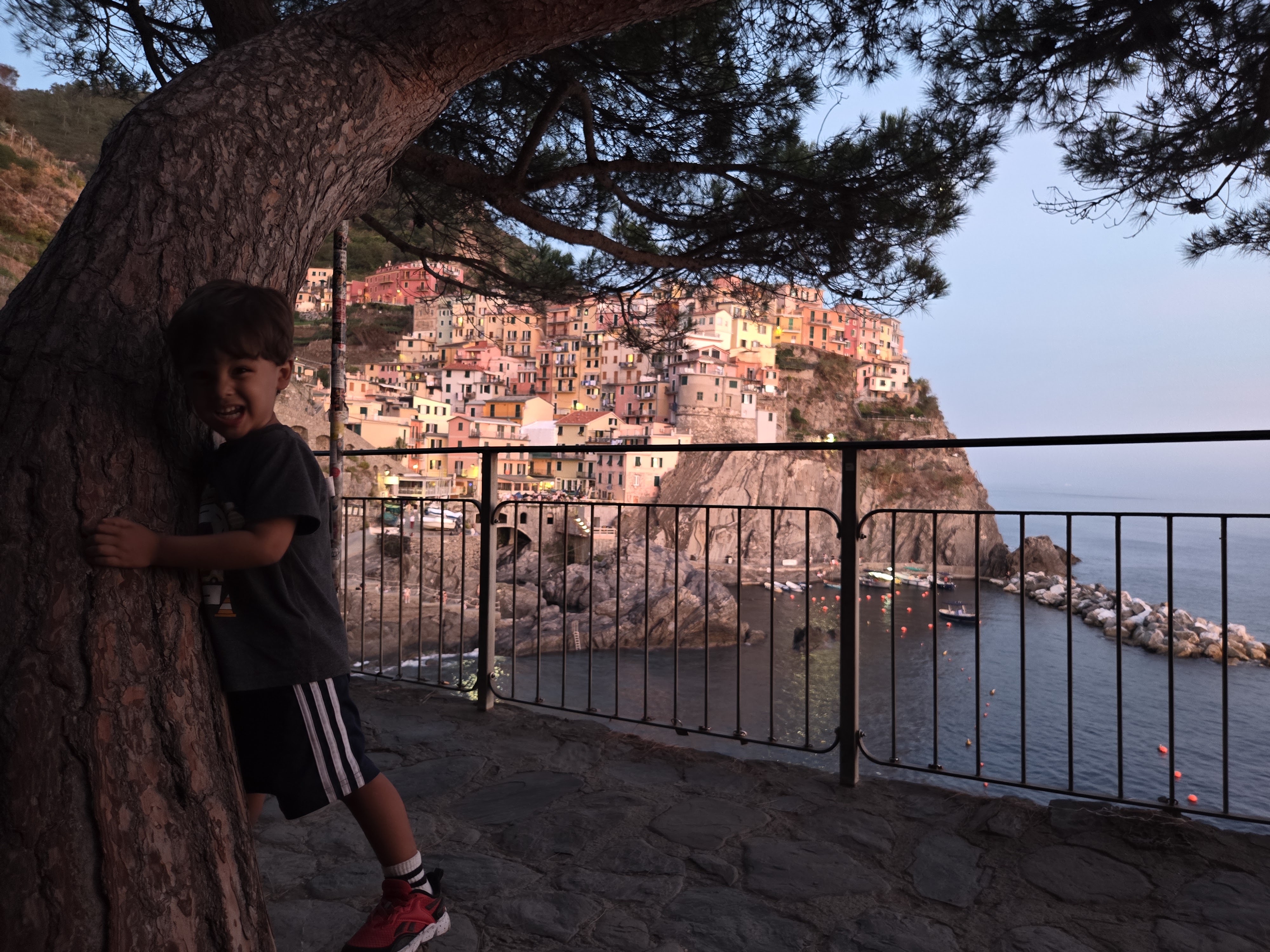Arlo leaning against a tree trunk, laughing, with Manarola's village and harbor visible through the railing behind him at dusk