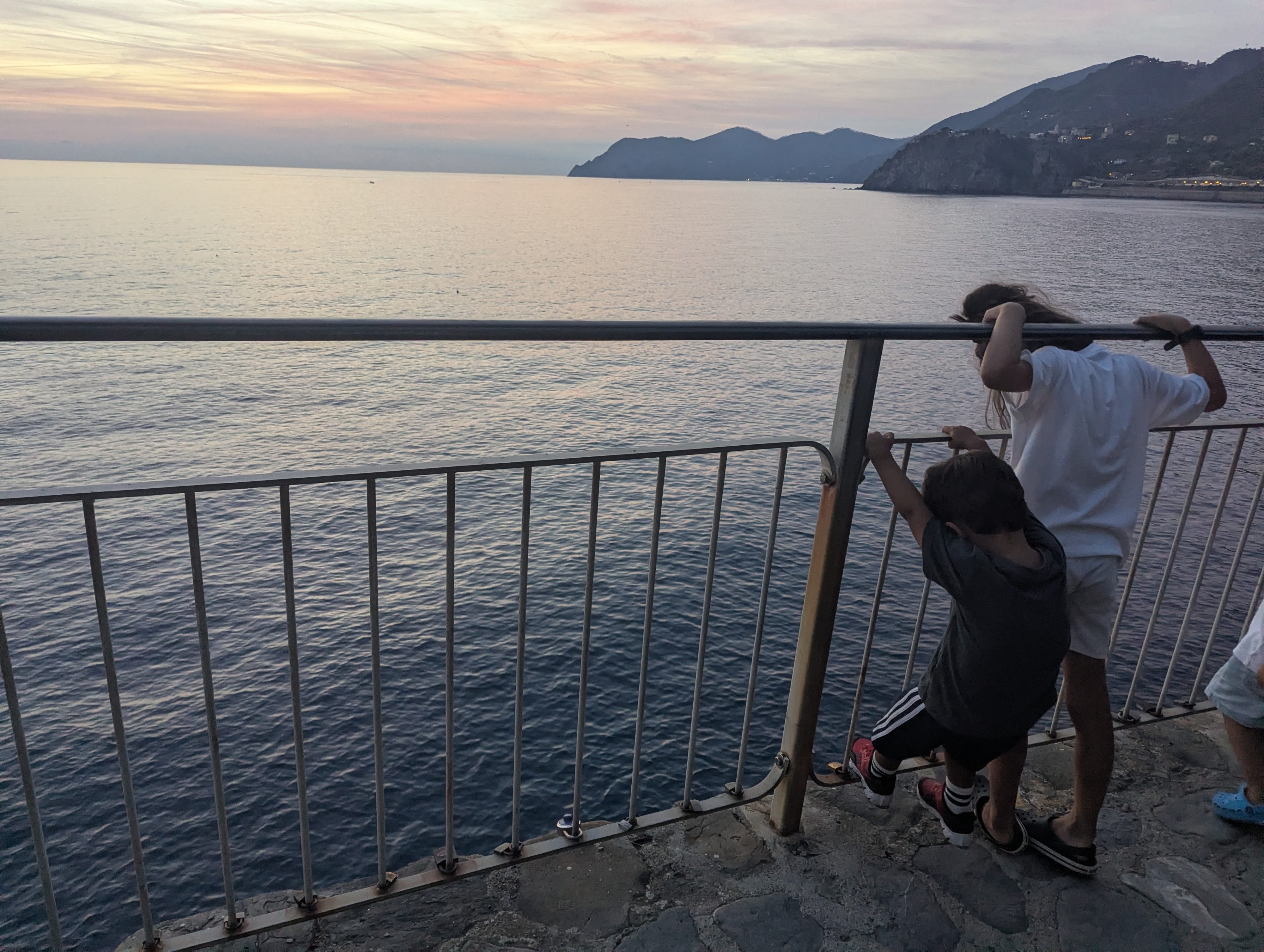 Remi and Arlo leaning over a metal railing above the sea at dusk, pink-streaked sky and the Ligurian headlands in the distance