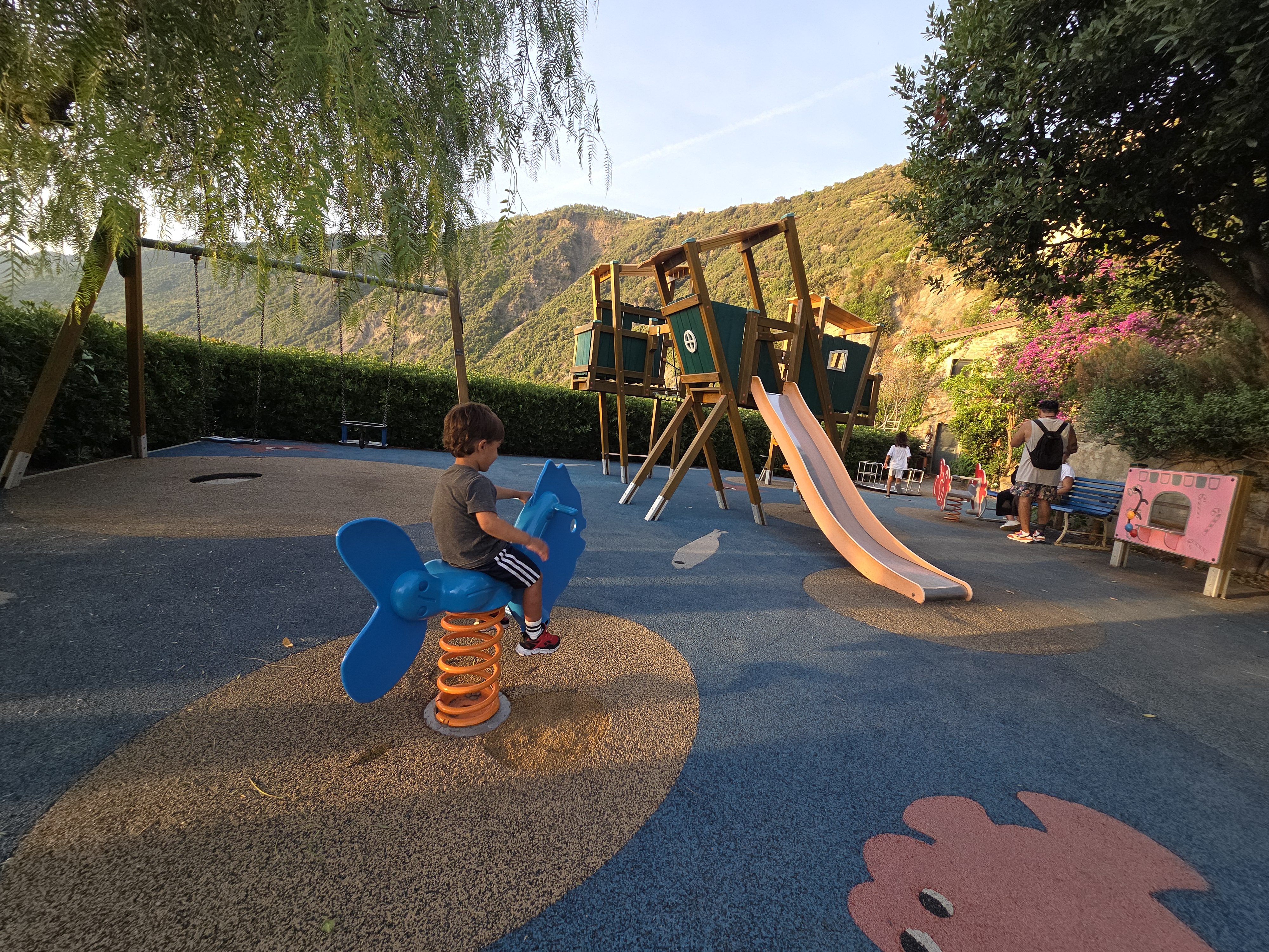 Arlo riding a blue spring fish at Manarola's playground, a wooden climbing frame with a slide and green hillside behind him