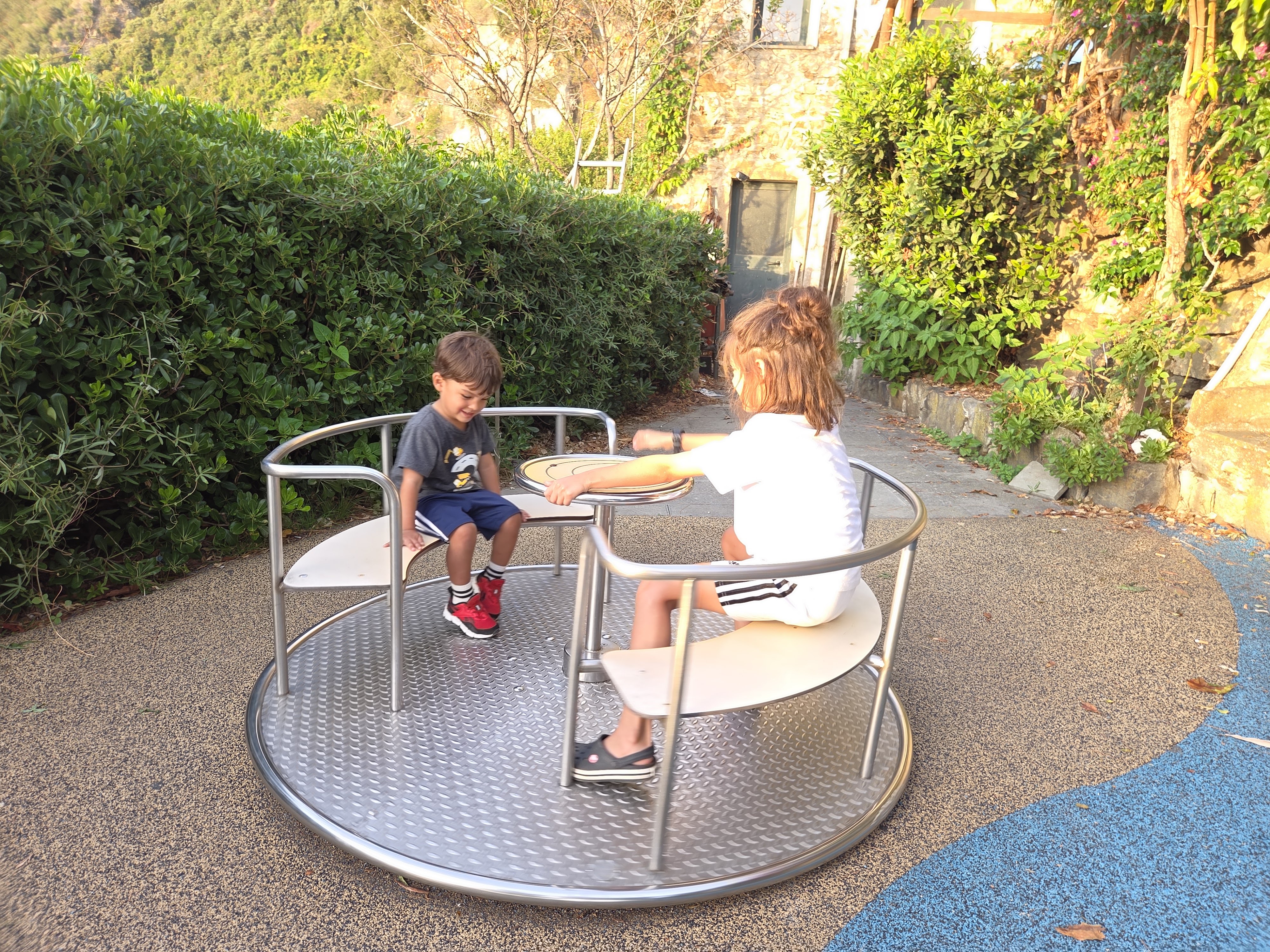 Two kids riding a metal roundabout at a small playground surrounded by greenery