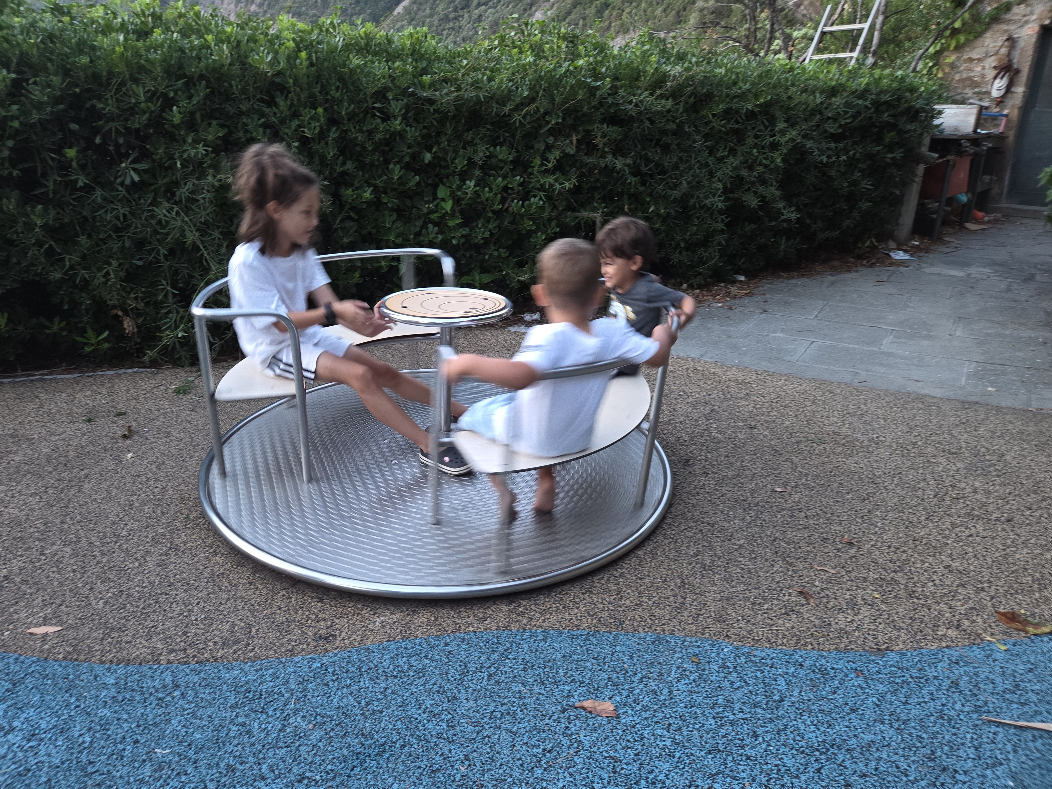 Remi, Arlo and another child spinning on a metal merry-go-round at the playground, motion-blurred