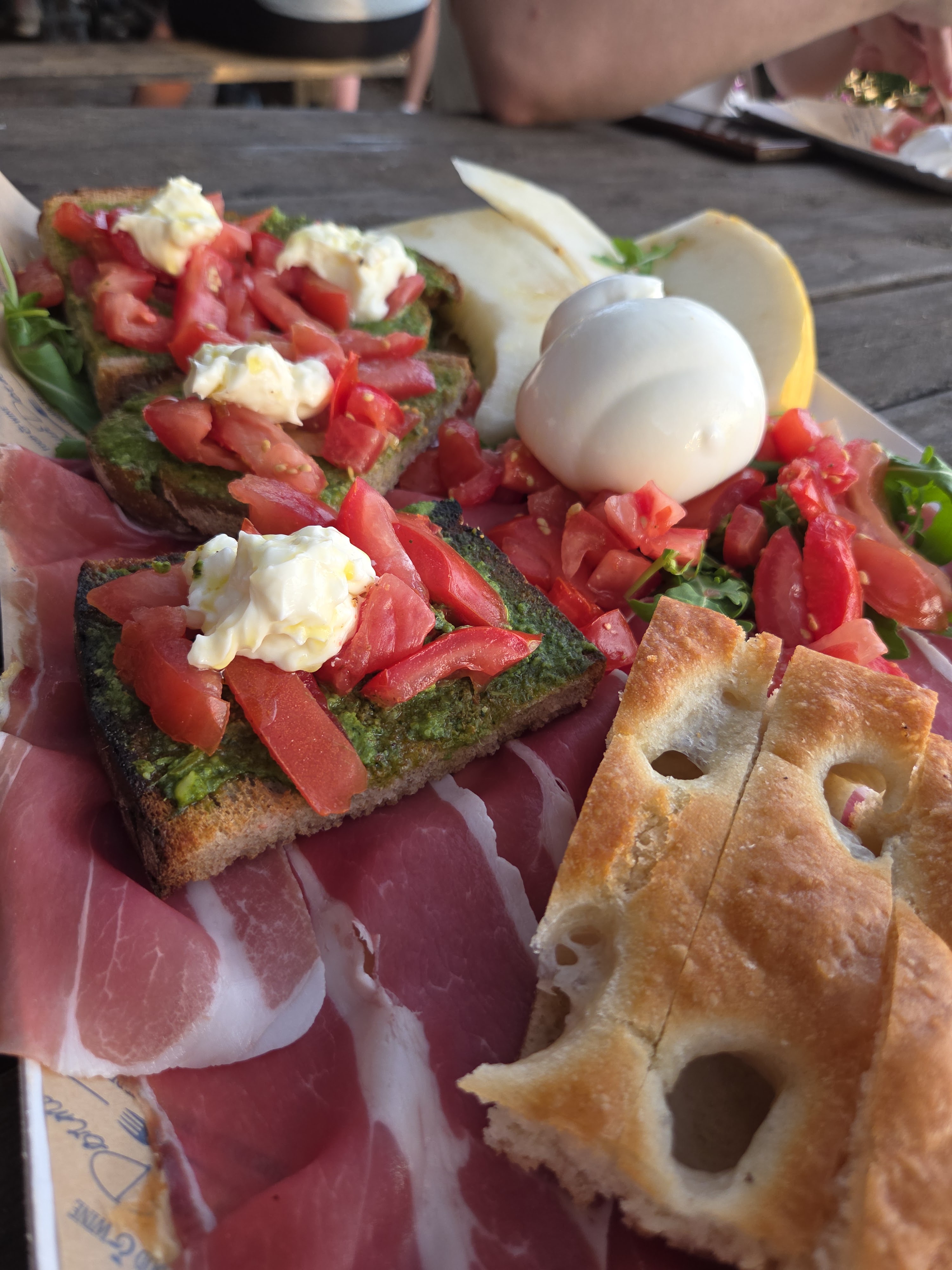 A to-go tasting spread of bruschetta with tomato and burrata, prosciutto, melon, and focaccia laid out on a wooden table
