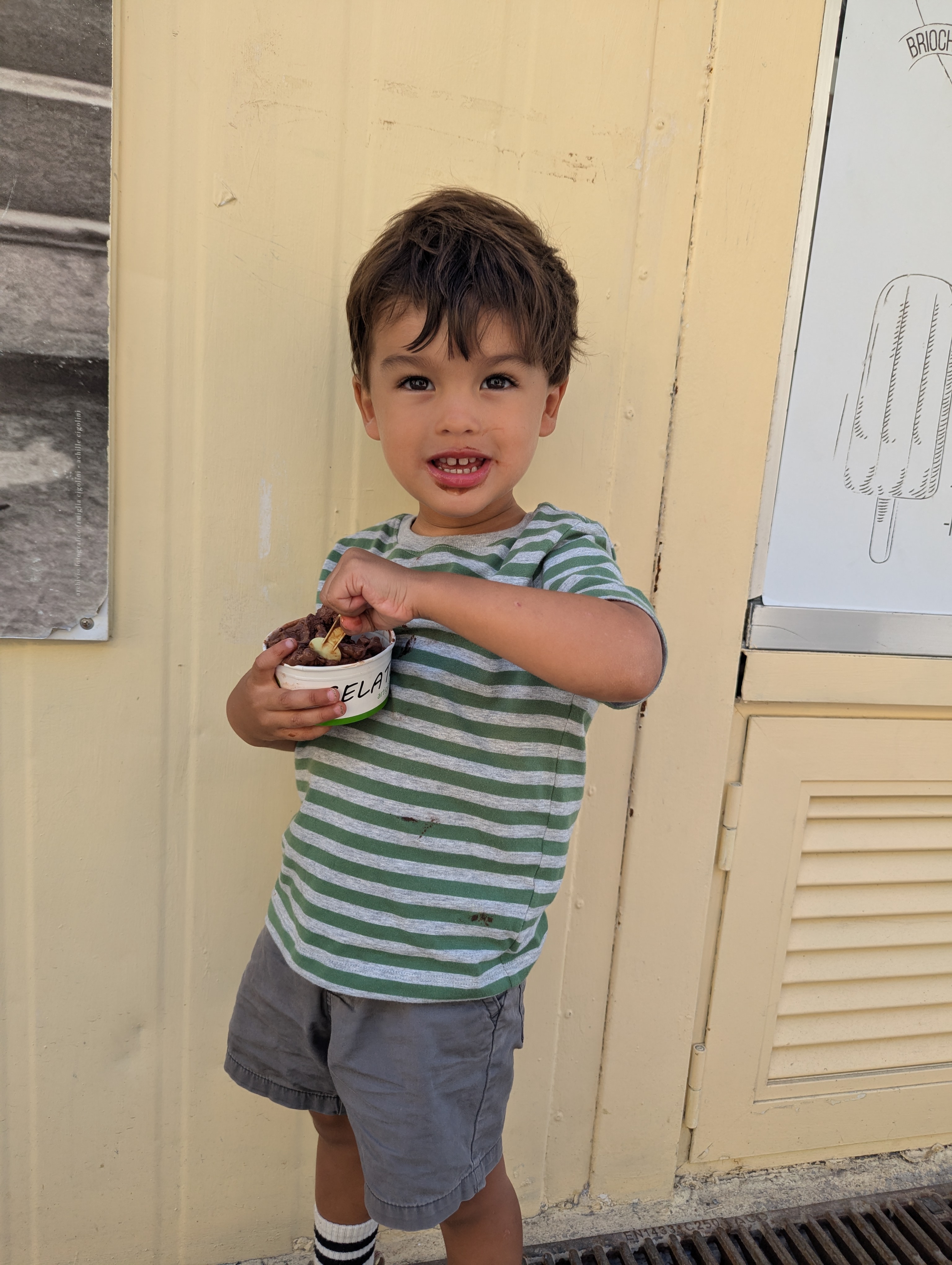 Arlo digging into a cup of chocolate gelato with a small wooden spoon, chocolate around his mouth