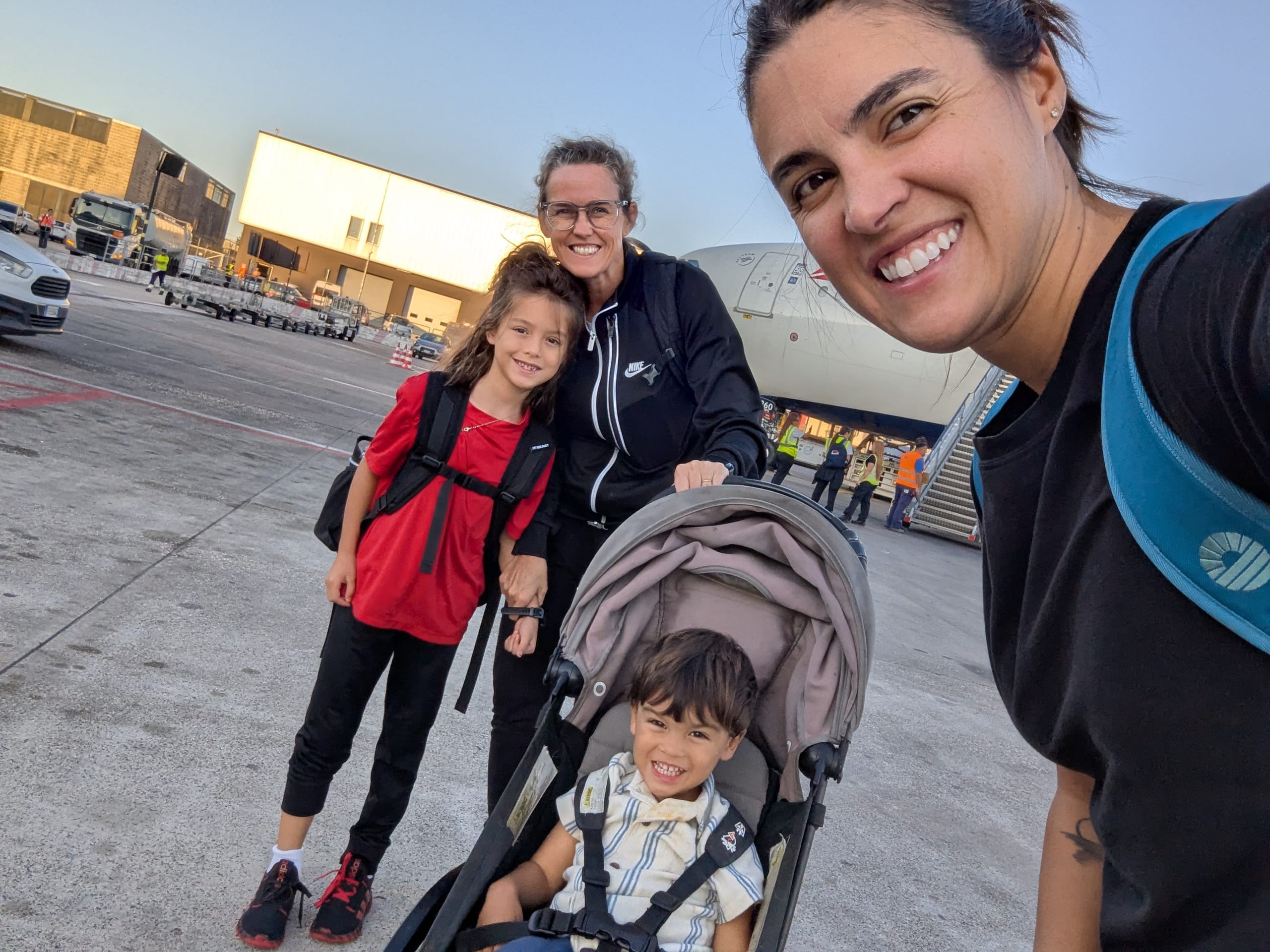 A selfie on the tarmac in Naples with the plane behind us, Remi in a red shirt with her backpack on and Arlo asleep in the stroller