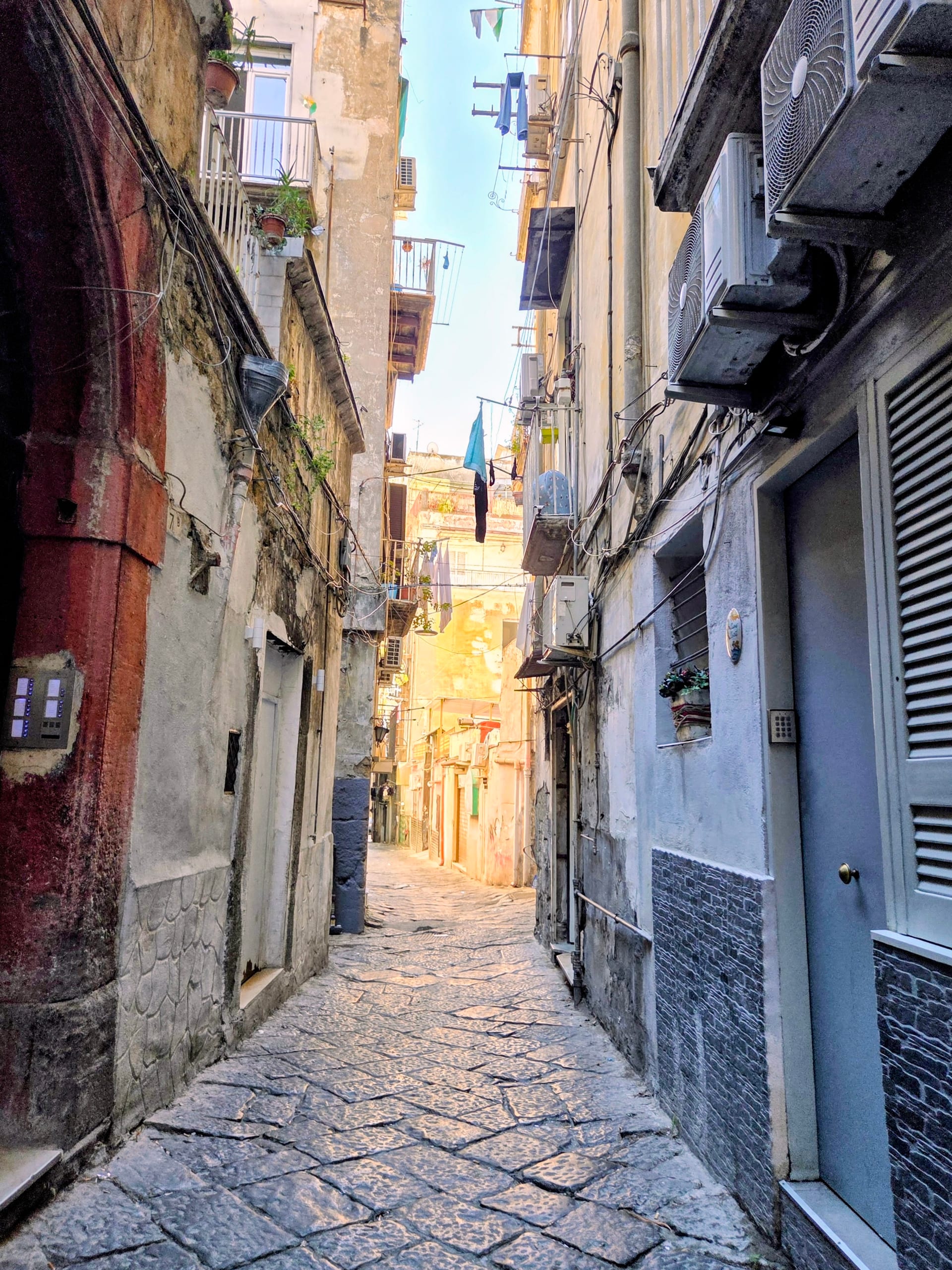 An empty cobblestone alley in the Naples historic center, balconies and laundry above and late sun running down the middle of the street