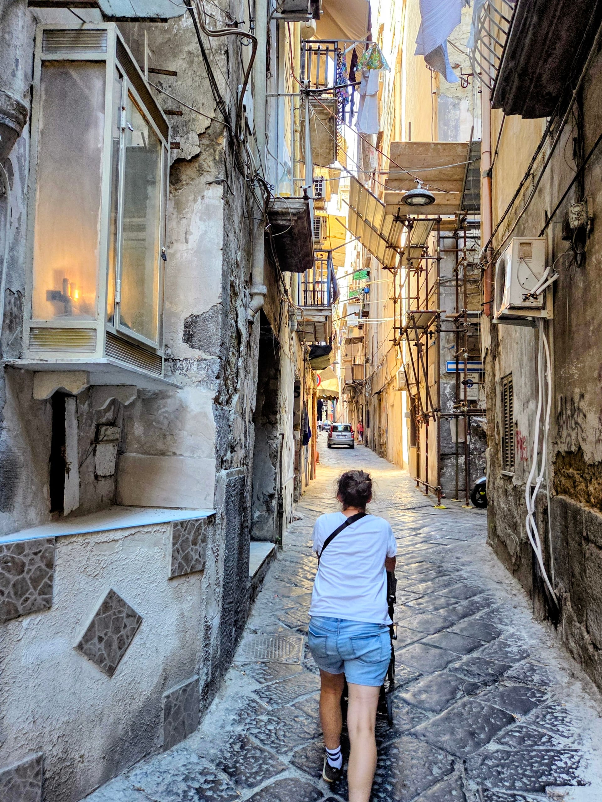 A narrow Naples alley of worn stone buildings with laundry strung overhead, the stroller being pushed down the cobblestones
