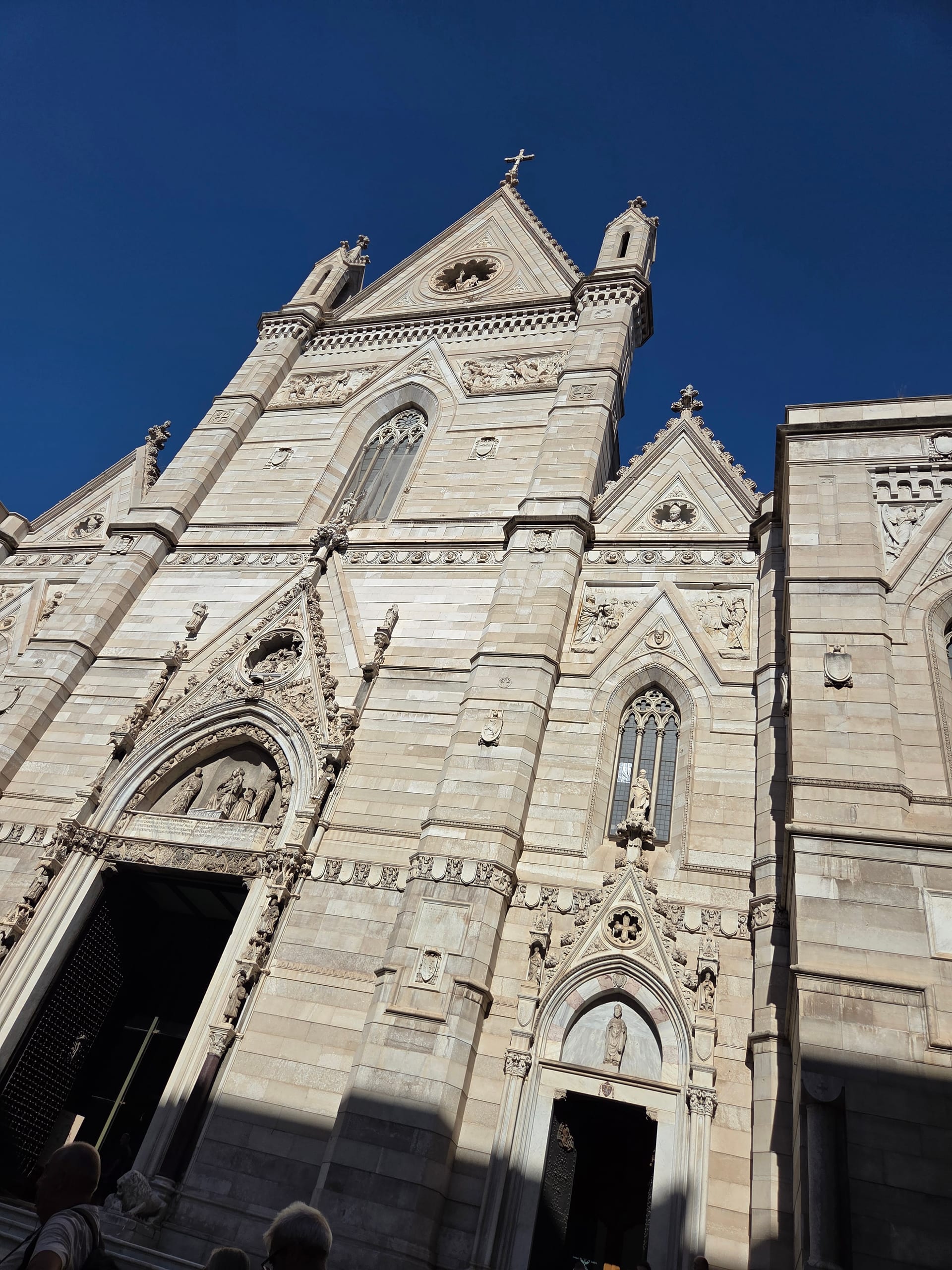 The carved stone facade of the Duomo di Napoli rising against a deep blue sky, photographed looking up from the steps