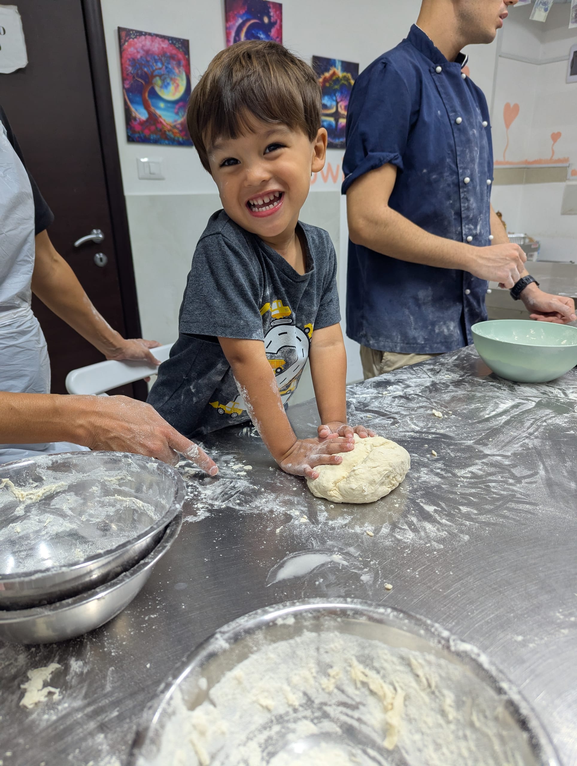 Arlo standing at the floured steel counter with both hands pressed into a ball of pizza dough, grinning at the camera