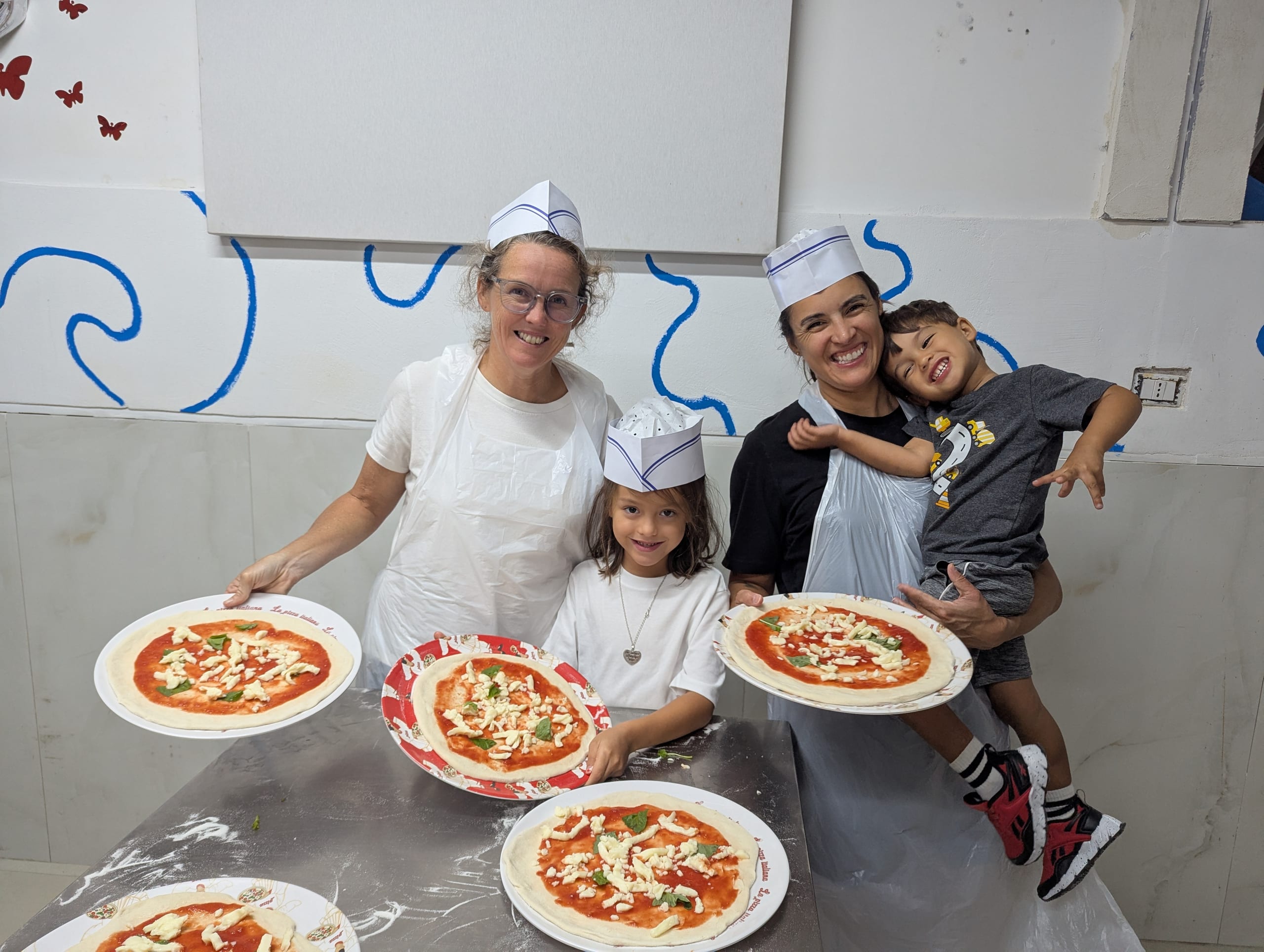 Alissa, Remi, Dani and Arlo in paper chef hats and aprons, each holding up the pizza they topped before it went in the oven