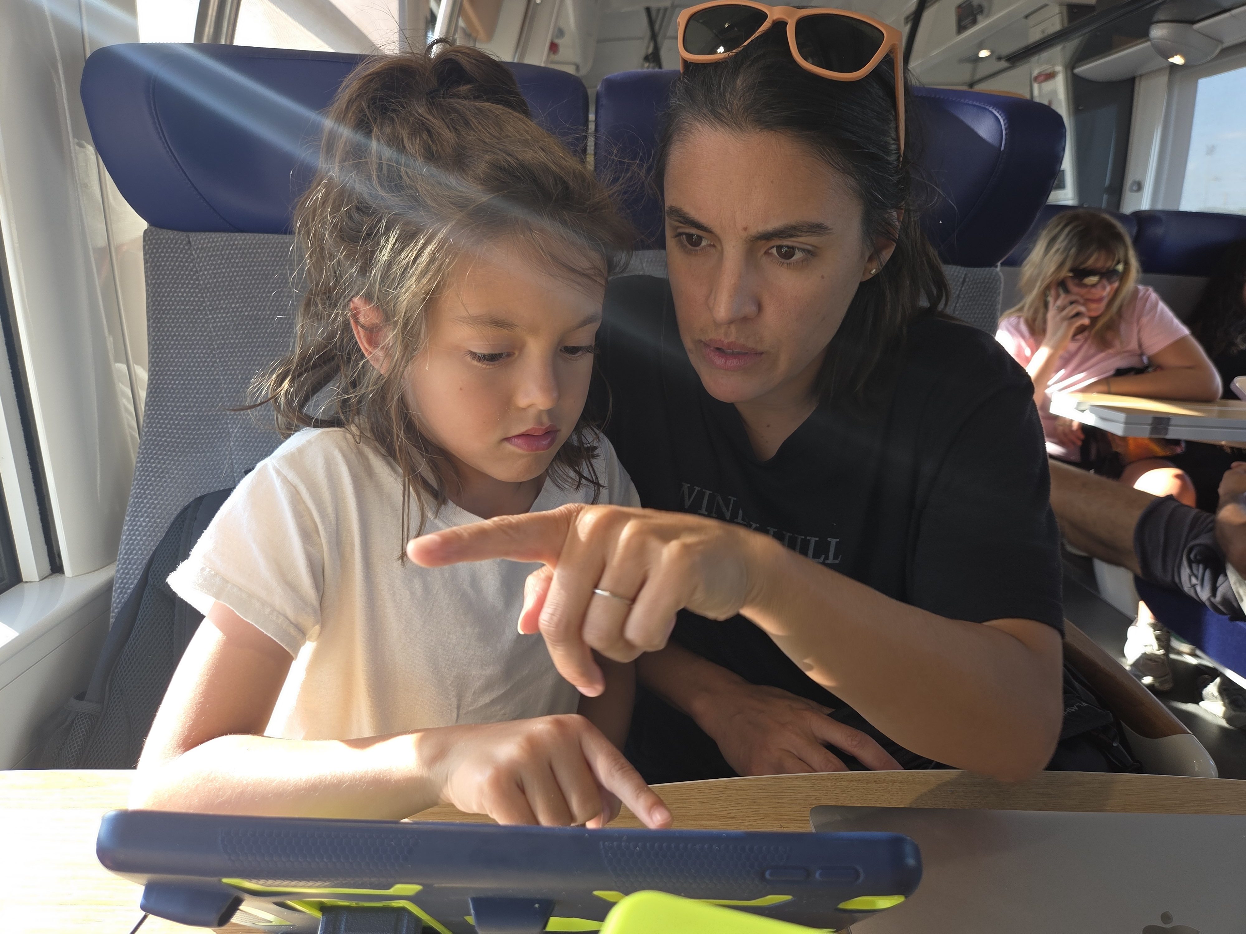 Alissa and Remi sitting together on a train, both looking at a tablet on the fold-down tray, sunlight streaming through the window