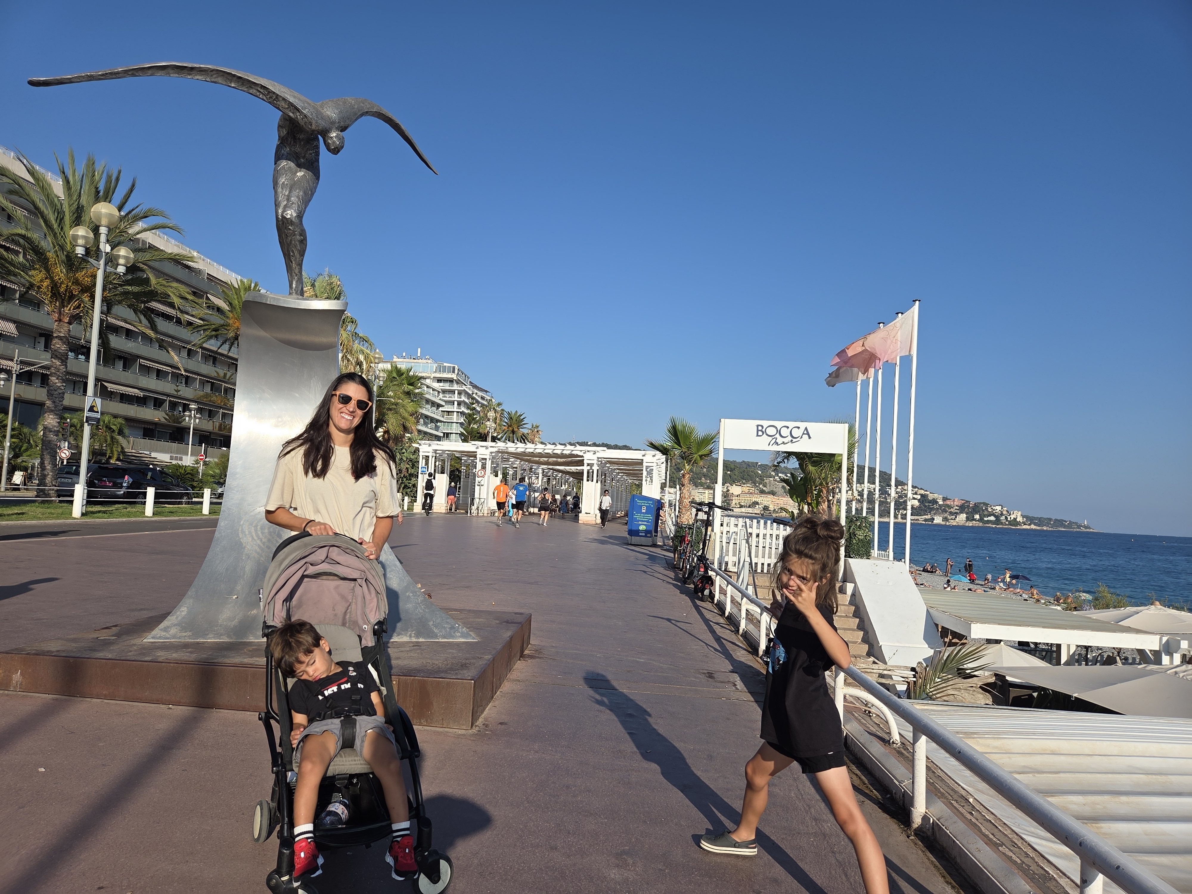 Alissa standing with Arlo in the stroller and Remi on the Promenade des Anglais in Nice, a diving sculpture and palm trees behind them, the beach and Mediterranean to the right
