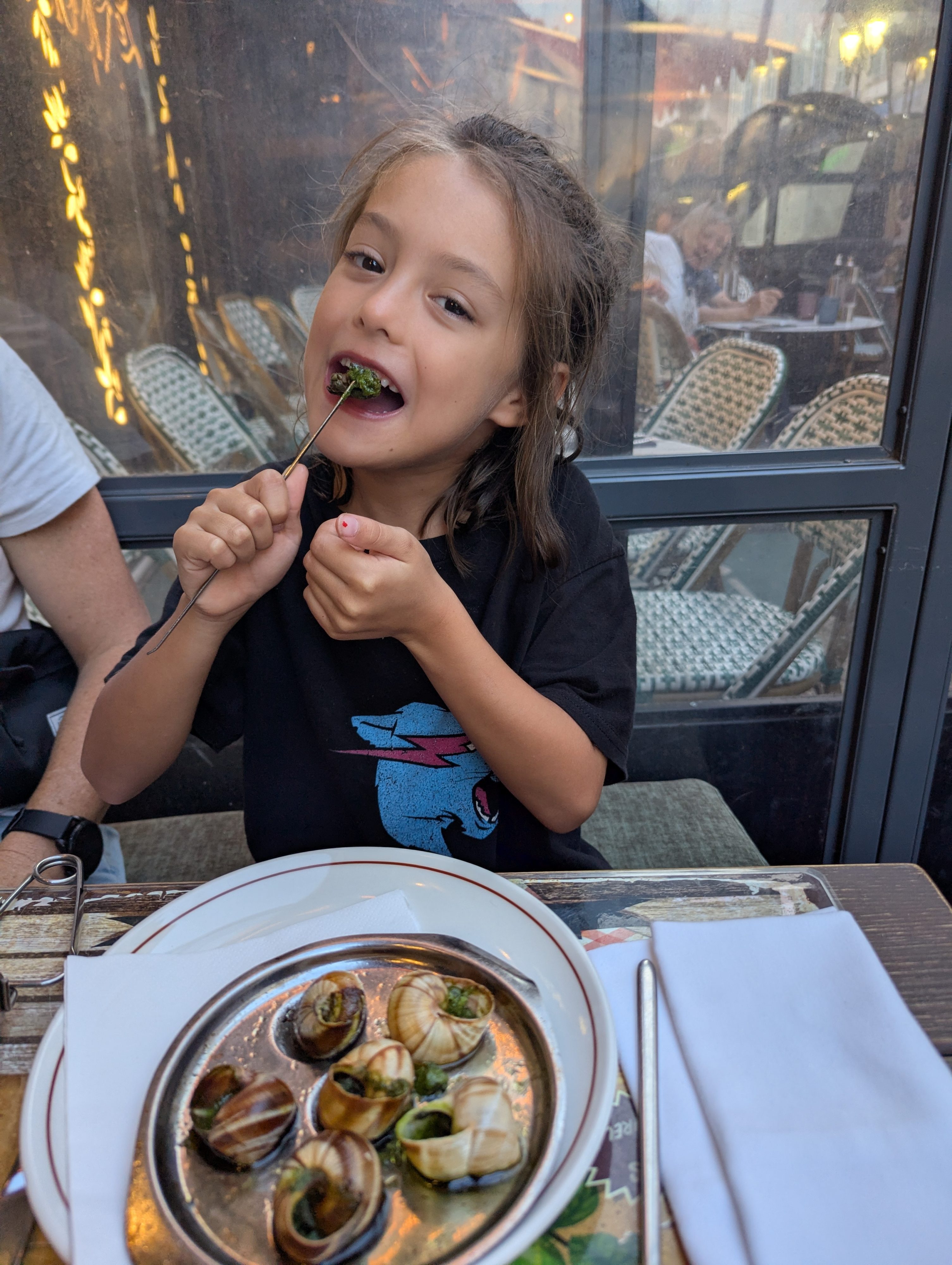 Remi holding an escargot on a small fork and grinning at a restaurant in Nice, a plate of snails in garlic butter in front of her