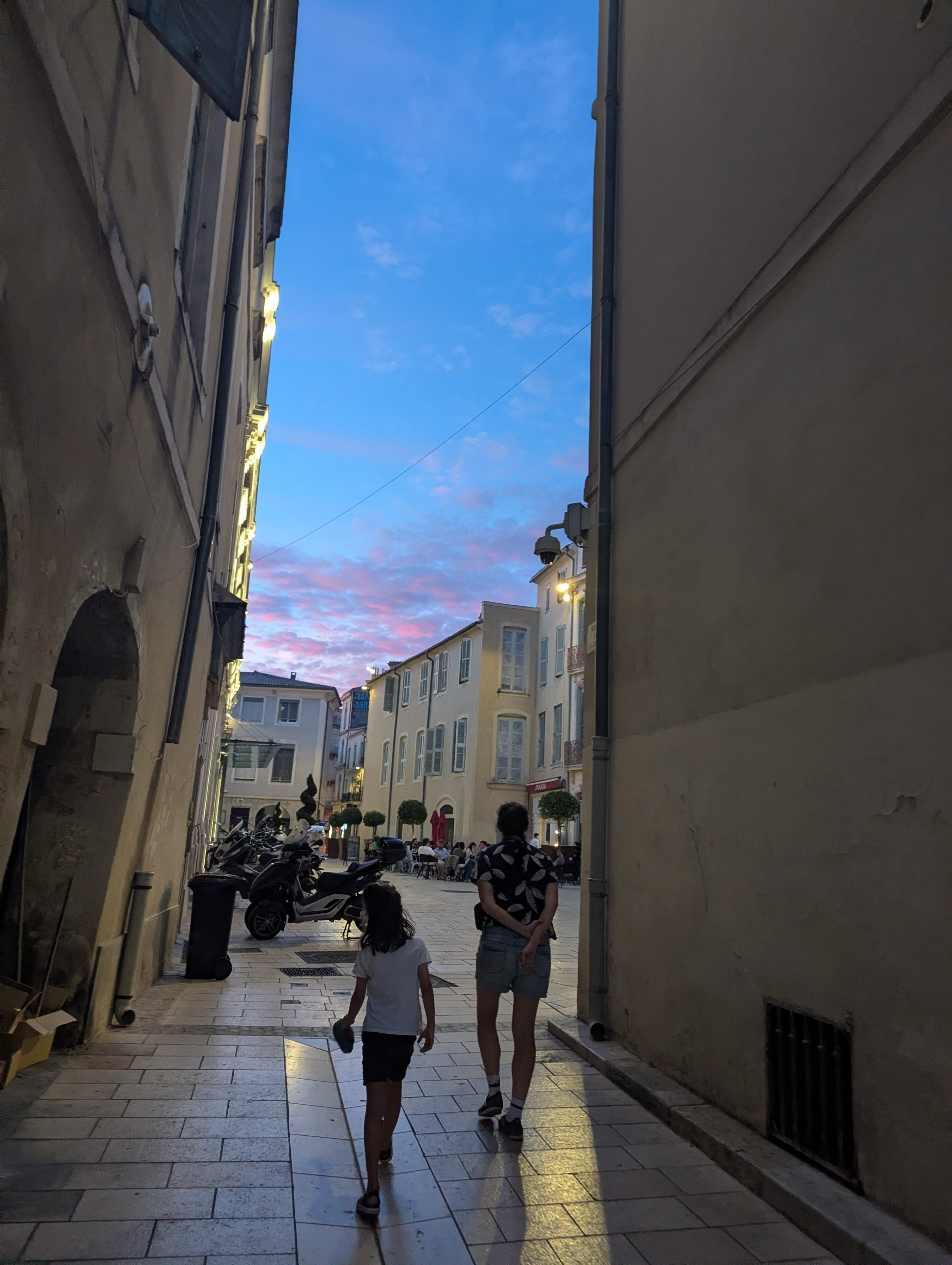 Dani and Remi walking through a narrow Nîmes alley toward a pink and blue sunset sky, silhouetted against the light at the far end