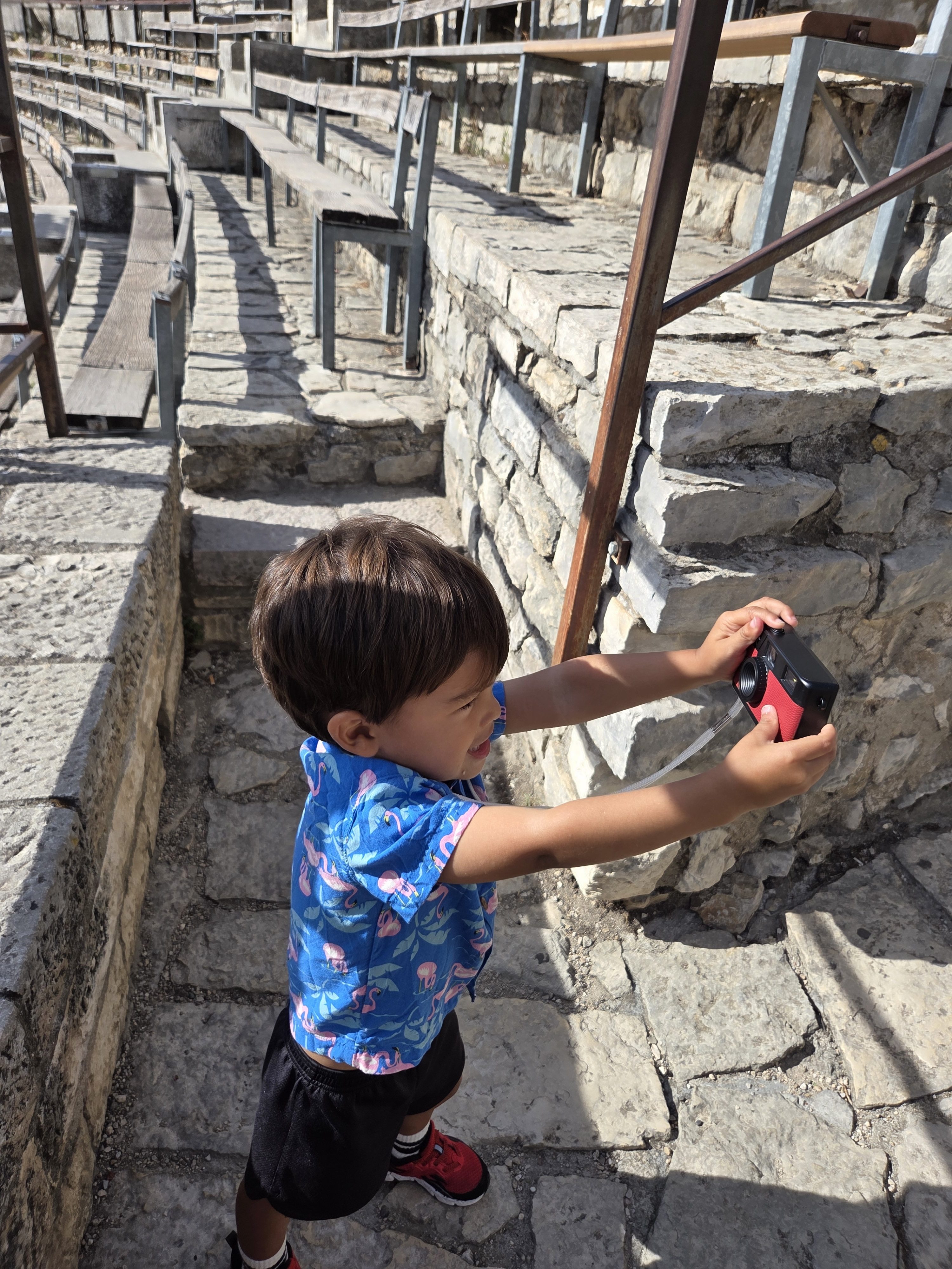 Arlo holding a small red camera up to the stone wall on the arena steps, concentrating hard on getting his shot