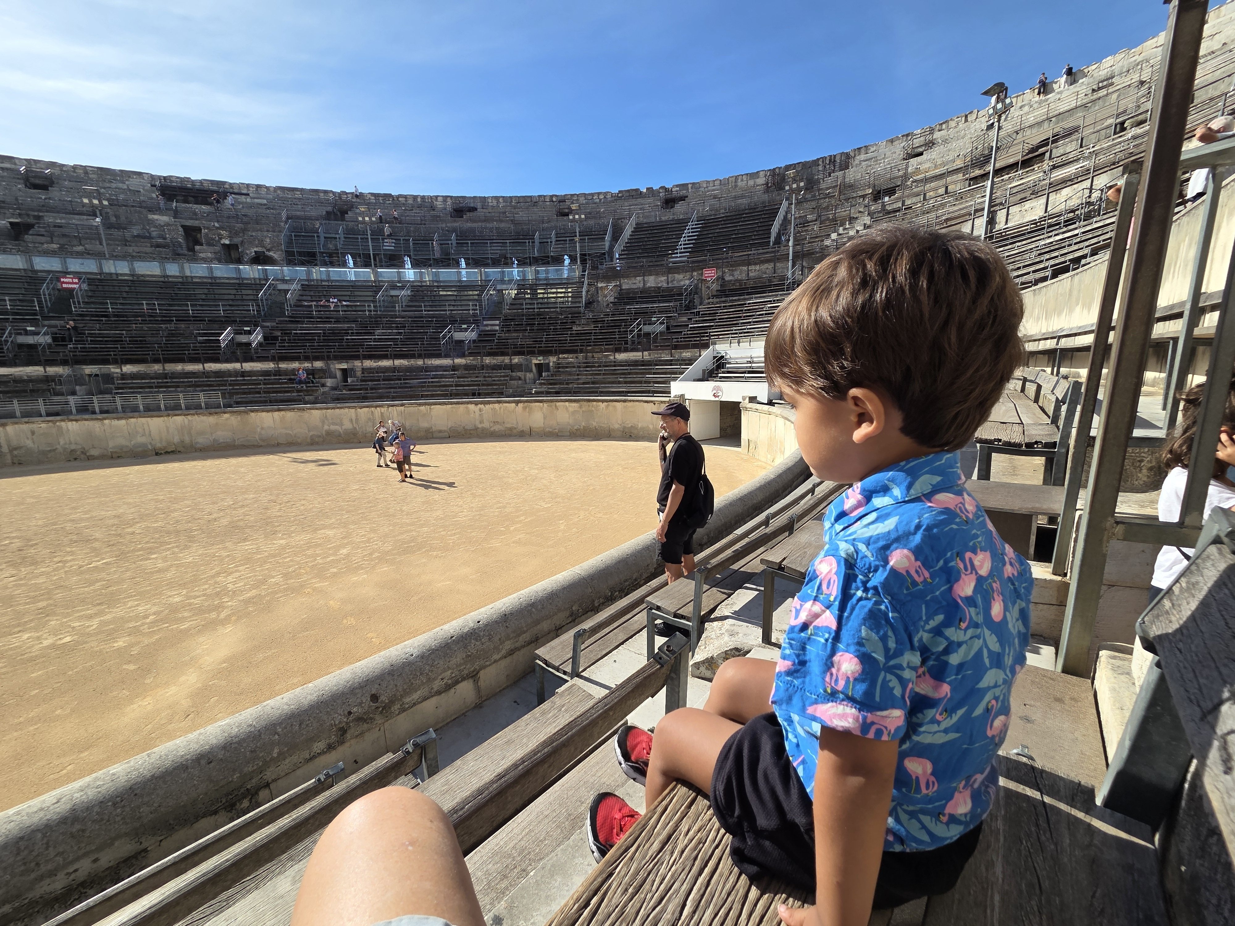 Arlo sitting on a wooden bench in the Arènes de Nîmes looking out at the sandy arena floor, with tiers of stone seating curving up behind him