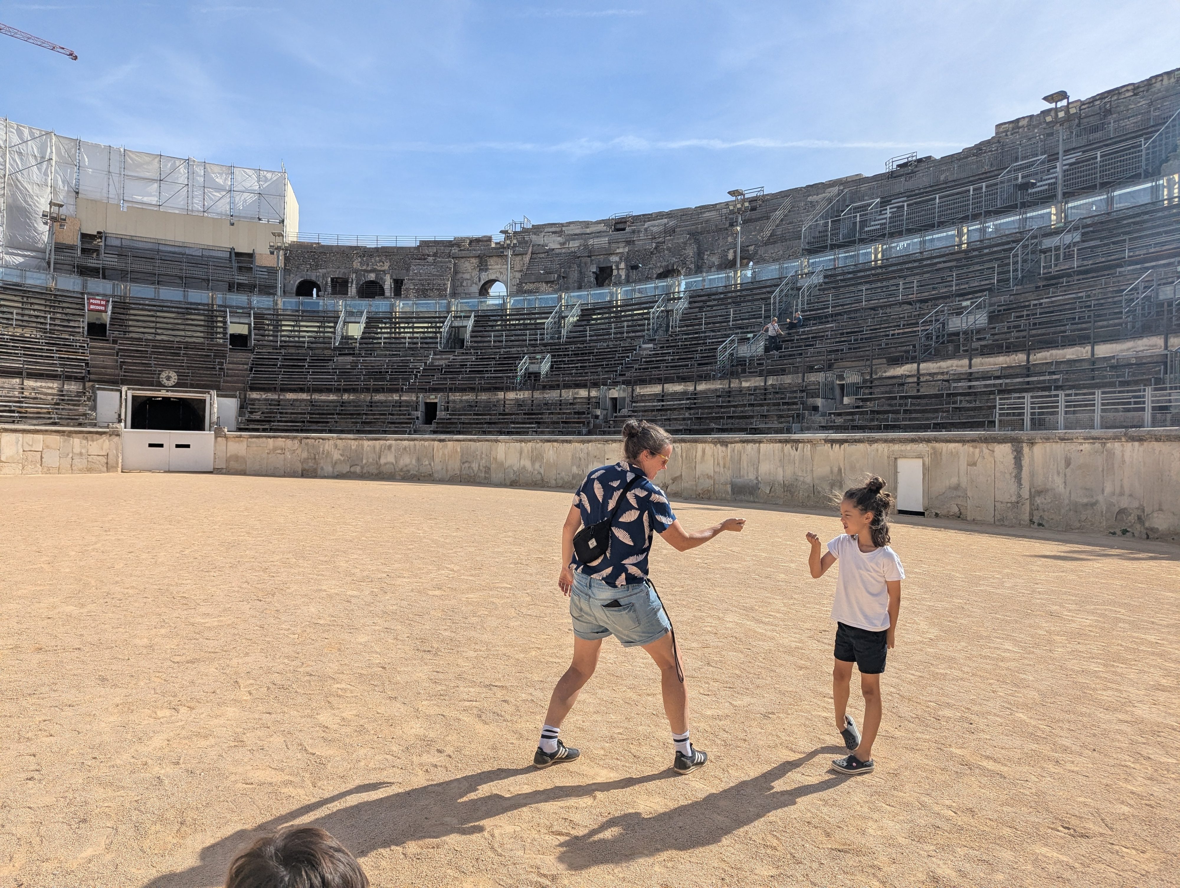 Dani and Remi facing each other in fighting stances on the sand floor of the Arènes de Nîmes, the curved stone tiers rising behind them