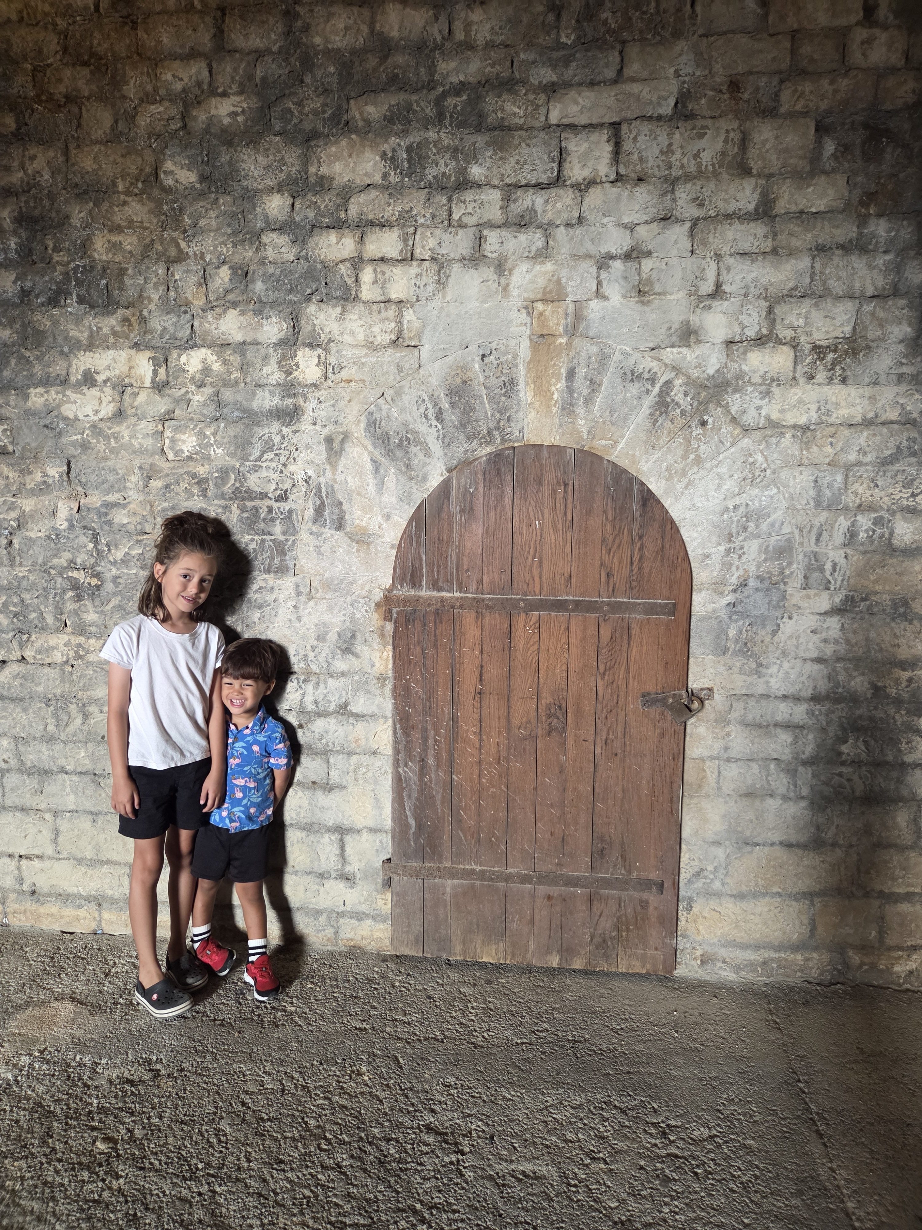 Remi and Arlo standing together in front of an arched wooden door set into the arena's stone wall, both smiling