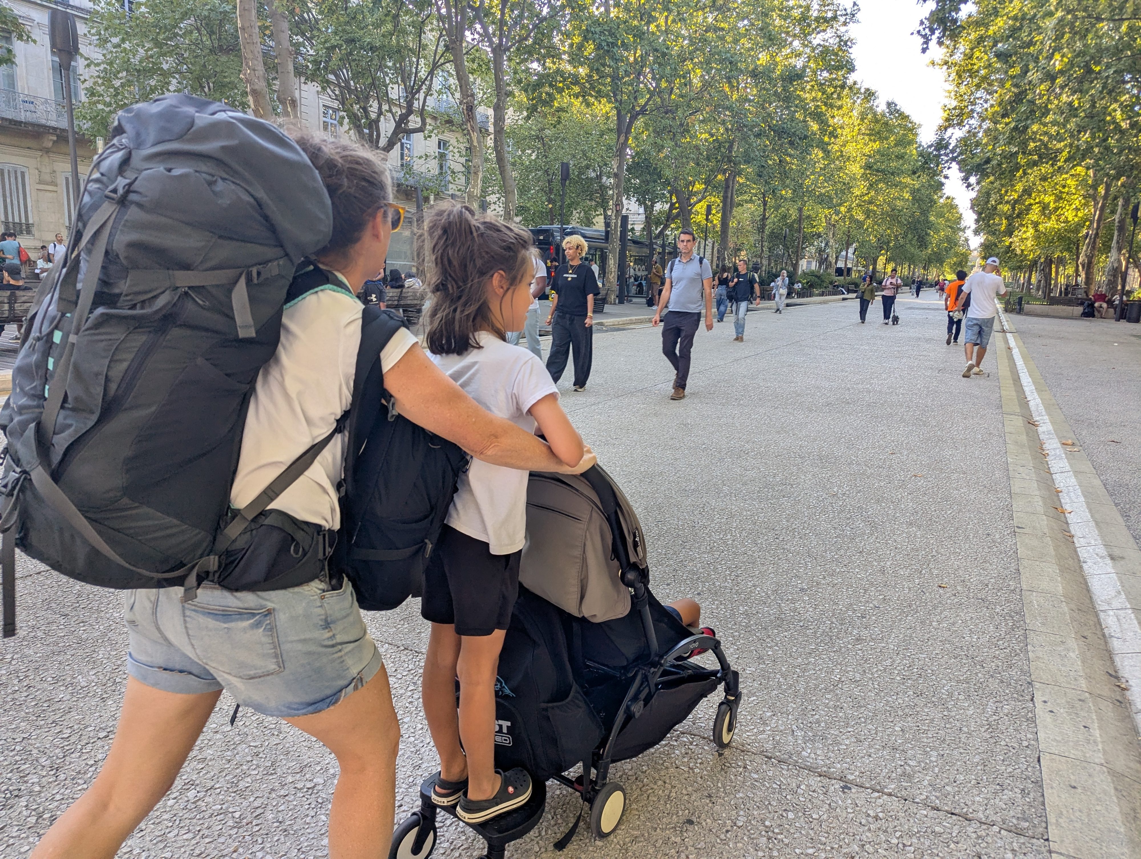 Dani carrying a full backpack and pushing the stroller with Remi walking alongside, heading down a wide tree-lined boulevard in Nîmes with other pedestrians around them