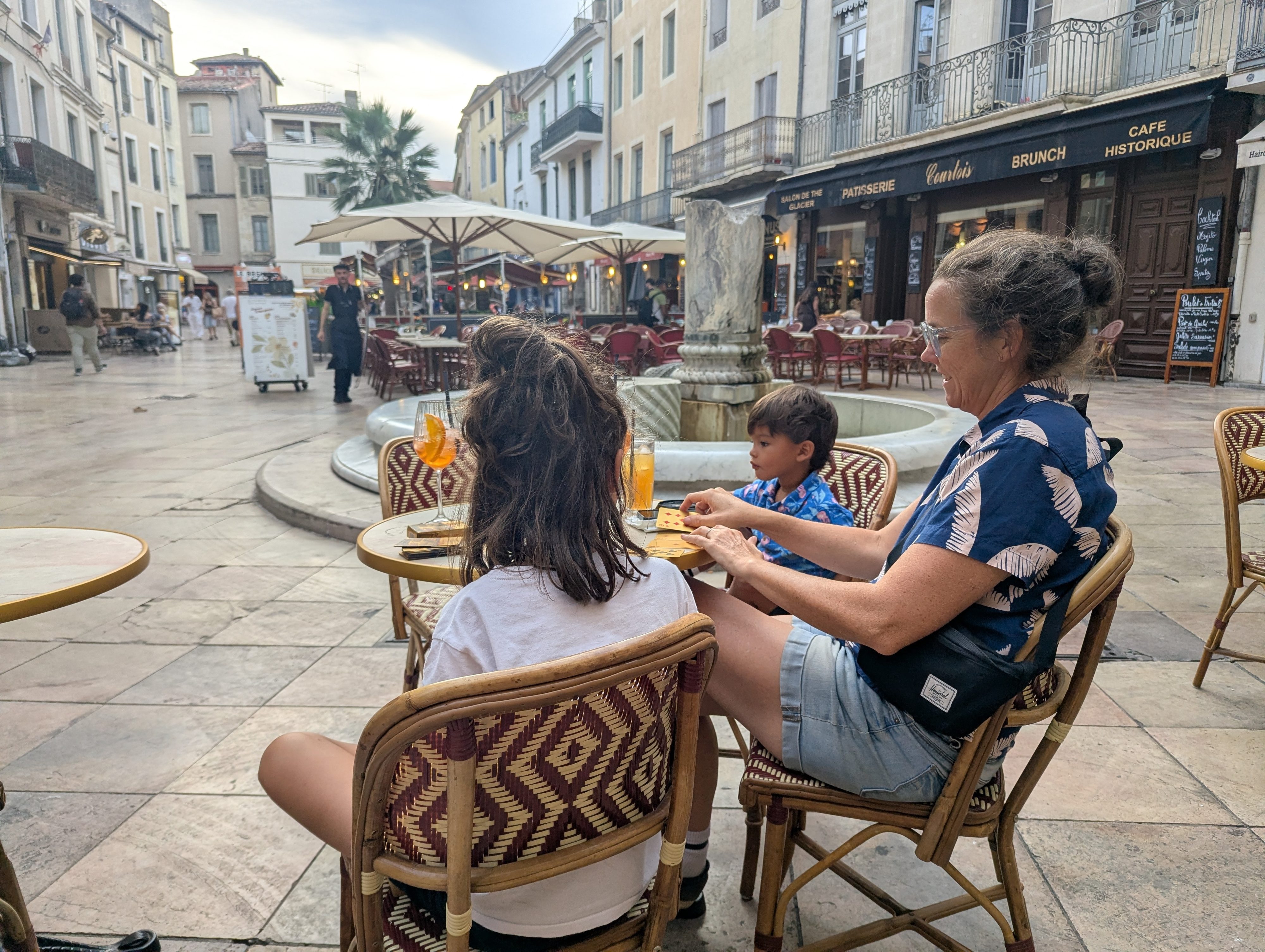 Dani sitting at a bistro table with Remi and Arlo in a Nîmes square at dusk, an Aperol spritz and juice on the table, a stone fountain and cafe patios in the background