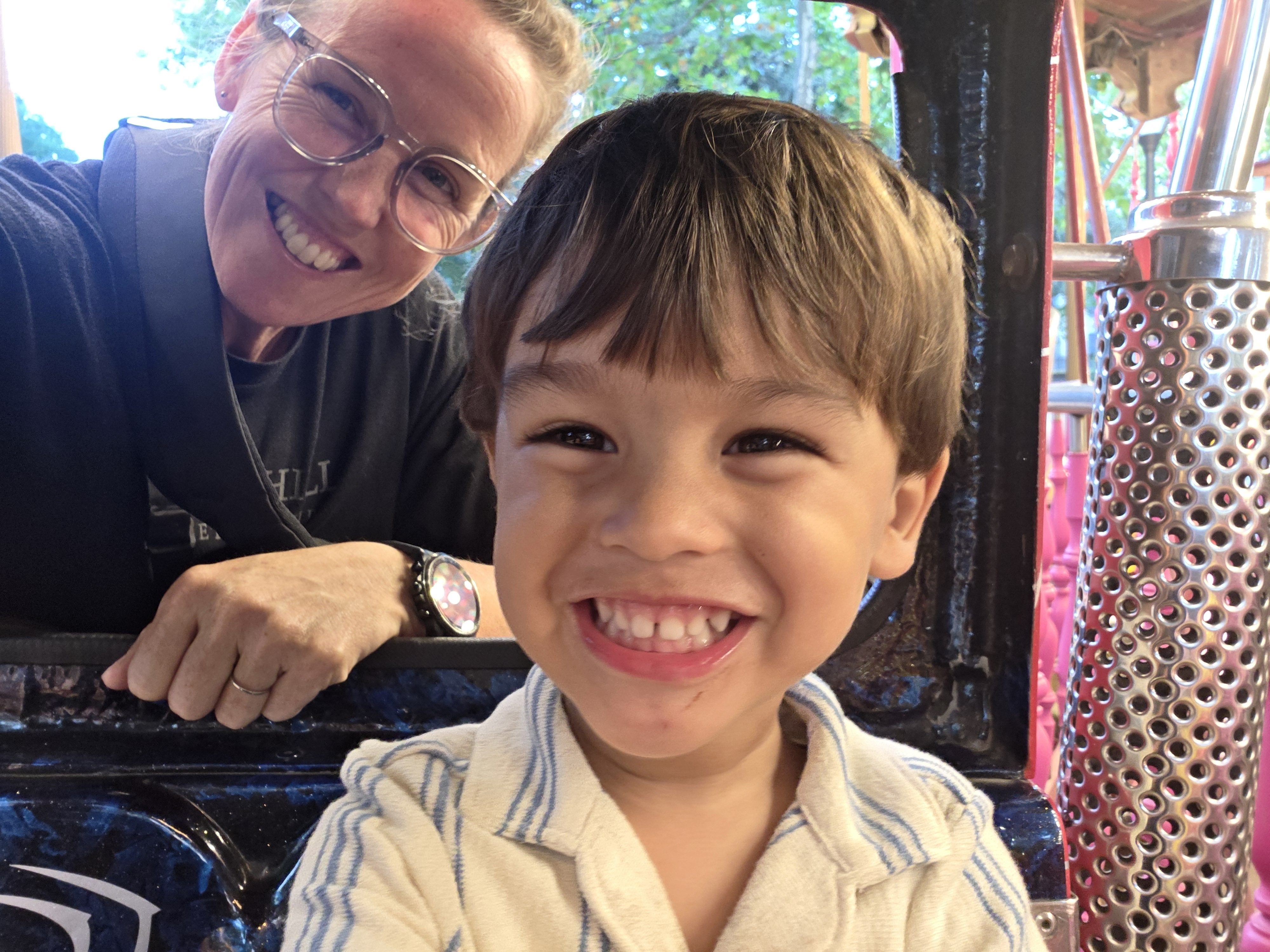 Arlo beaming at the camera from his seat on the carousel while a parent smiles over his shoulder
