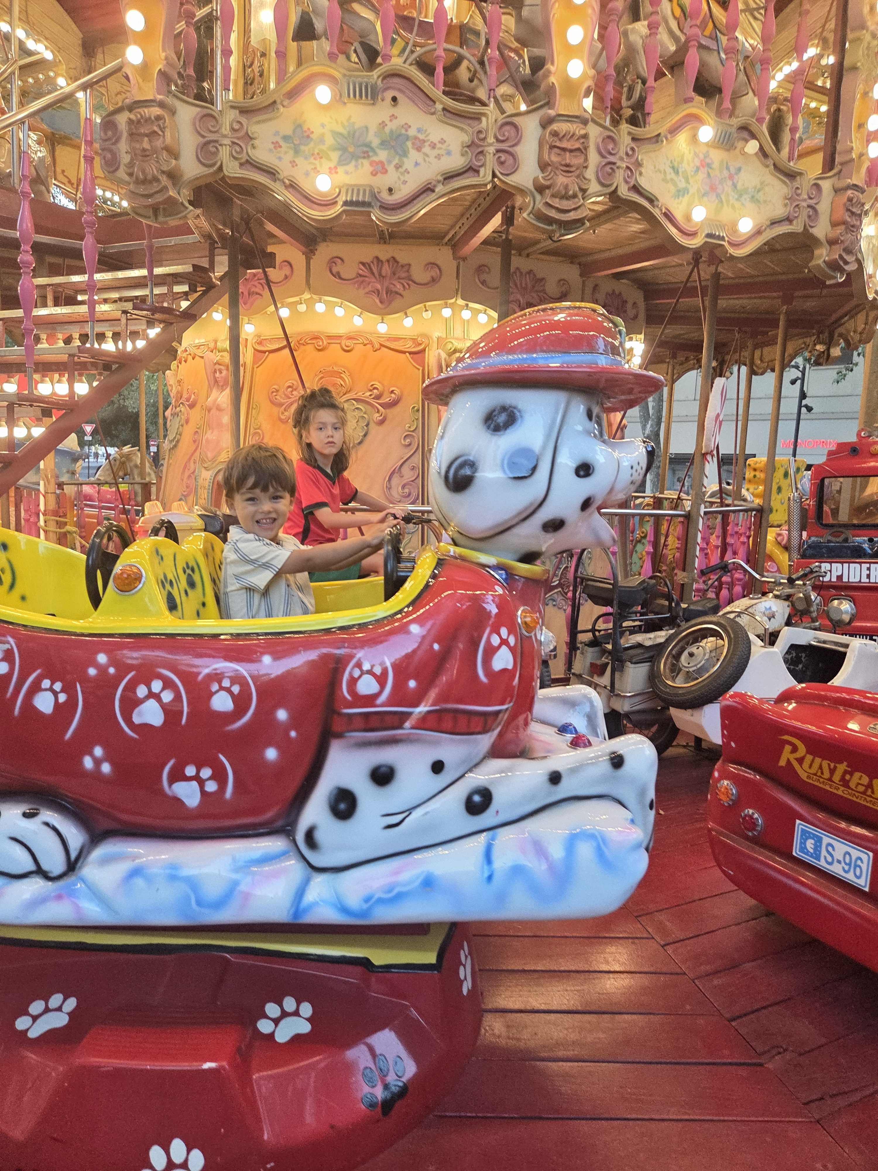 Arlo and Remi riding together in a red and yellow dalmatian fire-truck car on a lit-up, ornate carousel