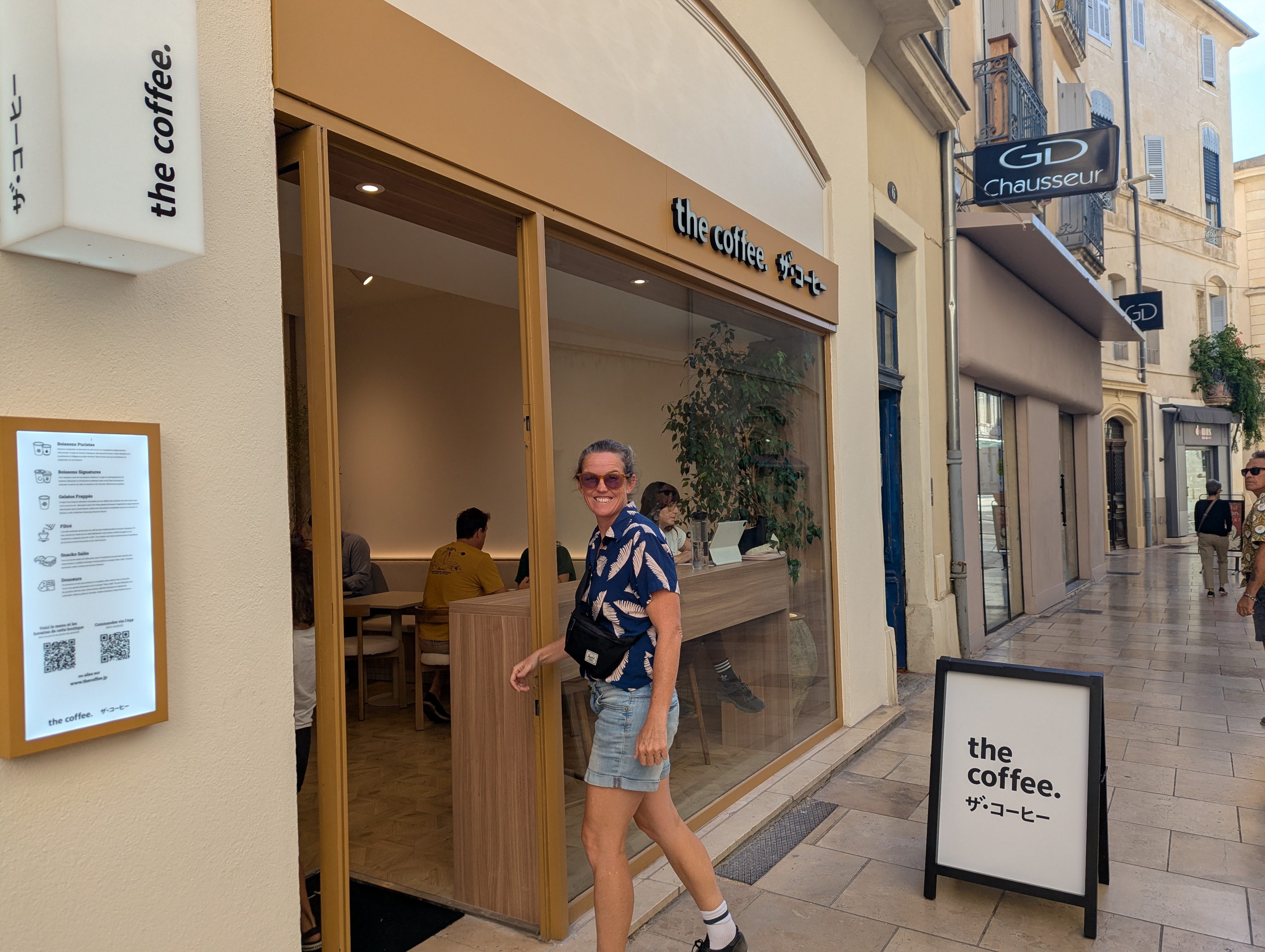Dani grinning as they walk into The Coffee, a minimalist Japanese-style coffee shop on a Nîmes pedestrian street, the menu board and sandwich sign flanking the open door
