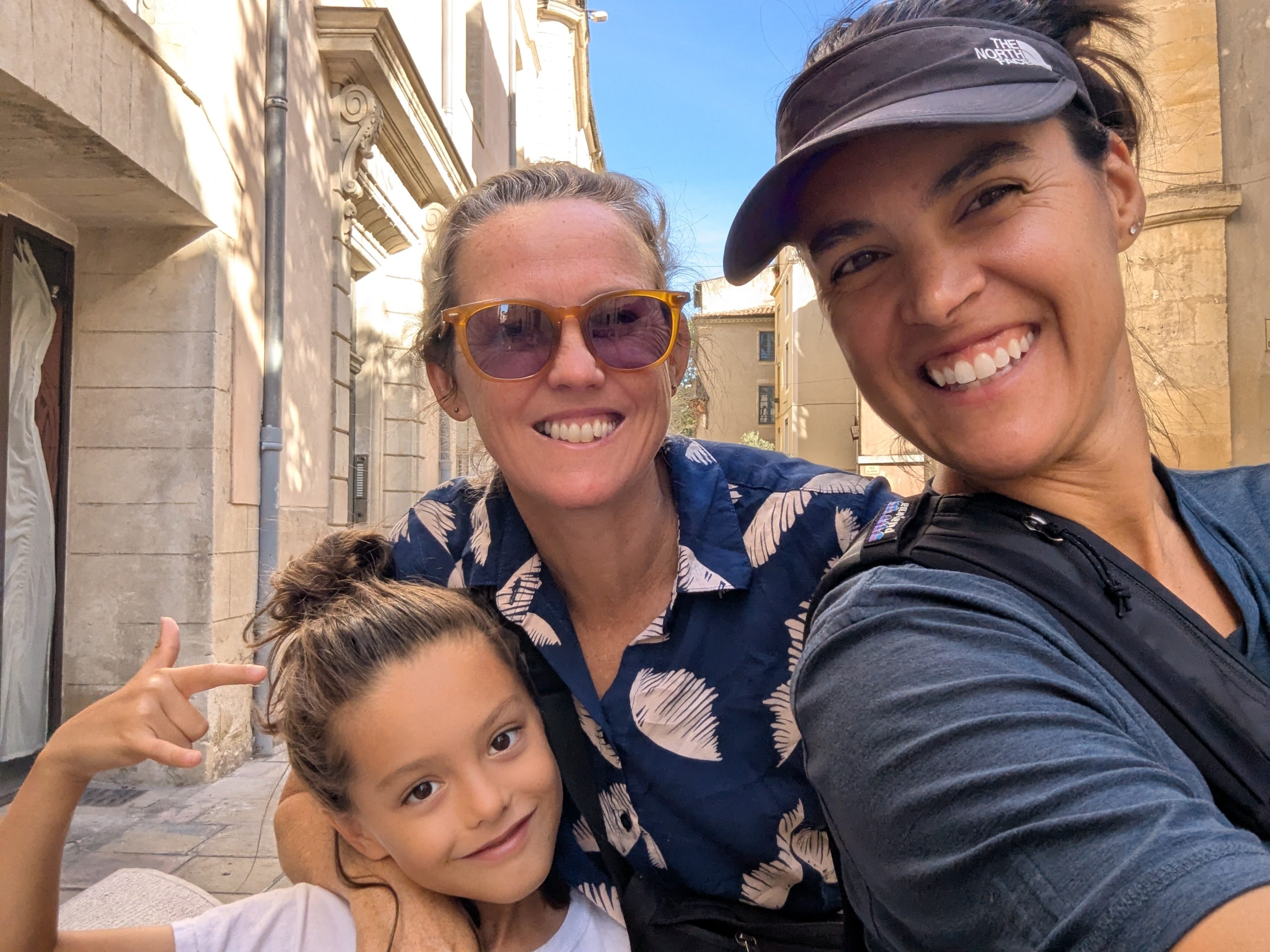 Alissa, Dani, and Remi in a selfie on a narrow Nîmes street, honey-colored stone buildings and blue sky behind them