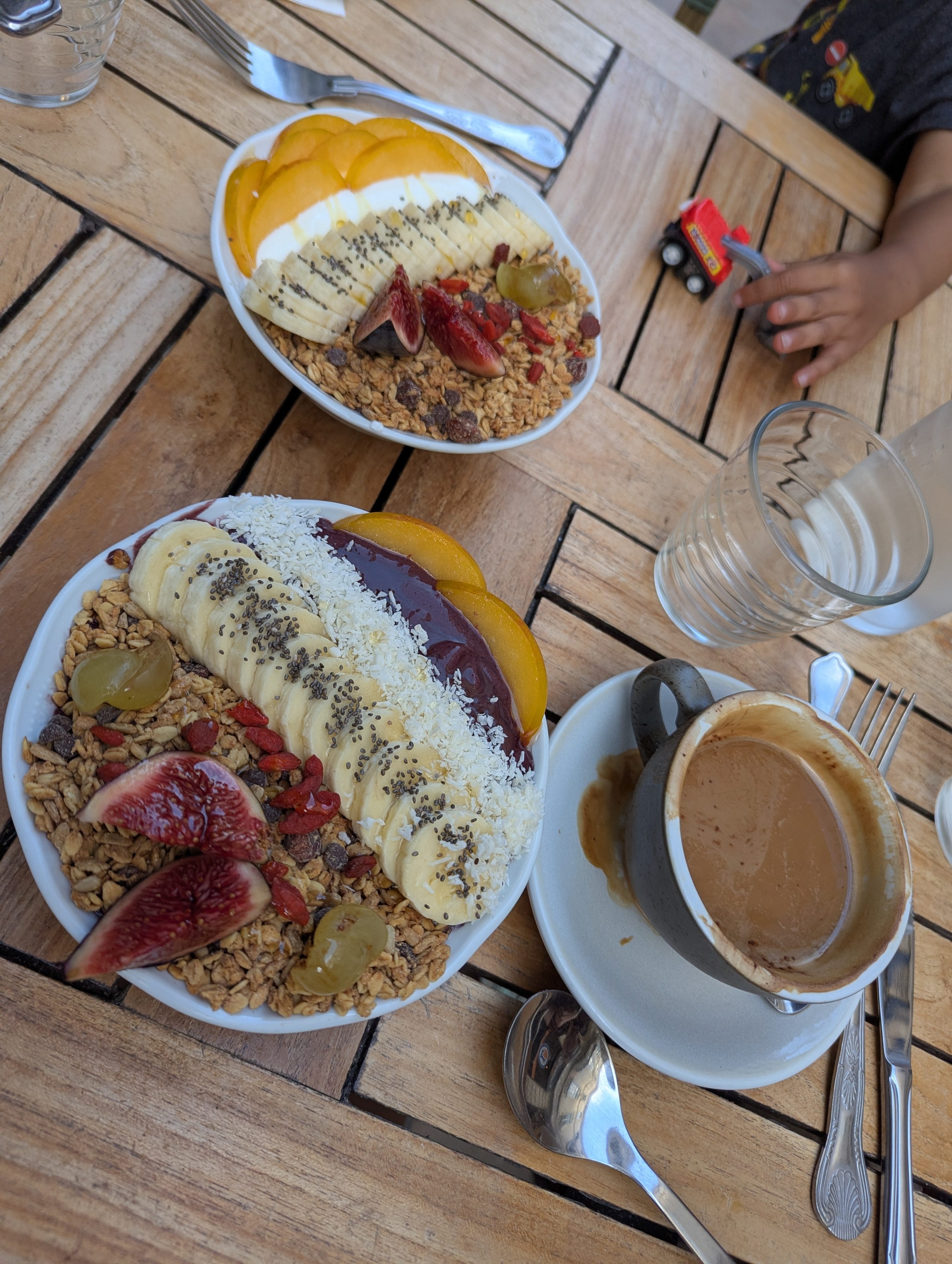 Two açaí and granola bowls topped with banana, fig, and coconut on a wooden table beside a coffee, with Arlo's hand pushing a small red toy truck at the edge of the frame