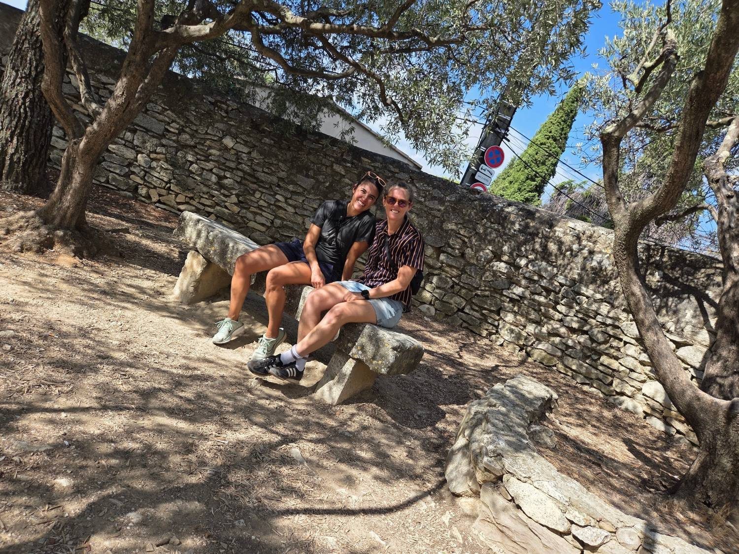 Two parents sitting close together and smiling on a worn stone bench in the dappled shade of olive trees, a dry-stone wall behind them