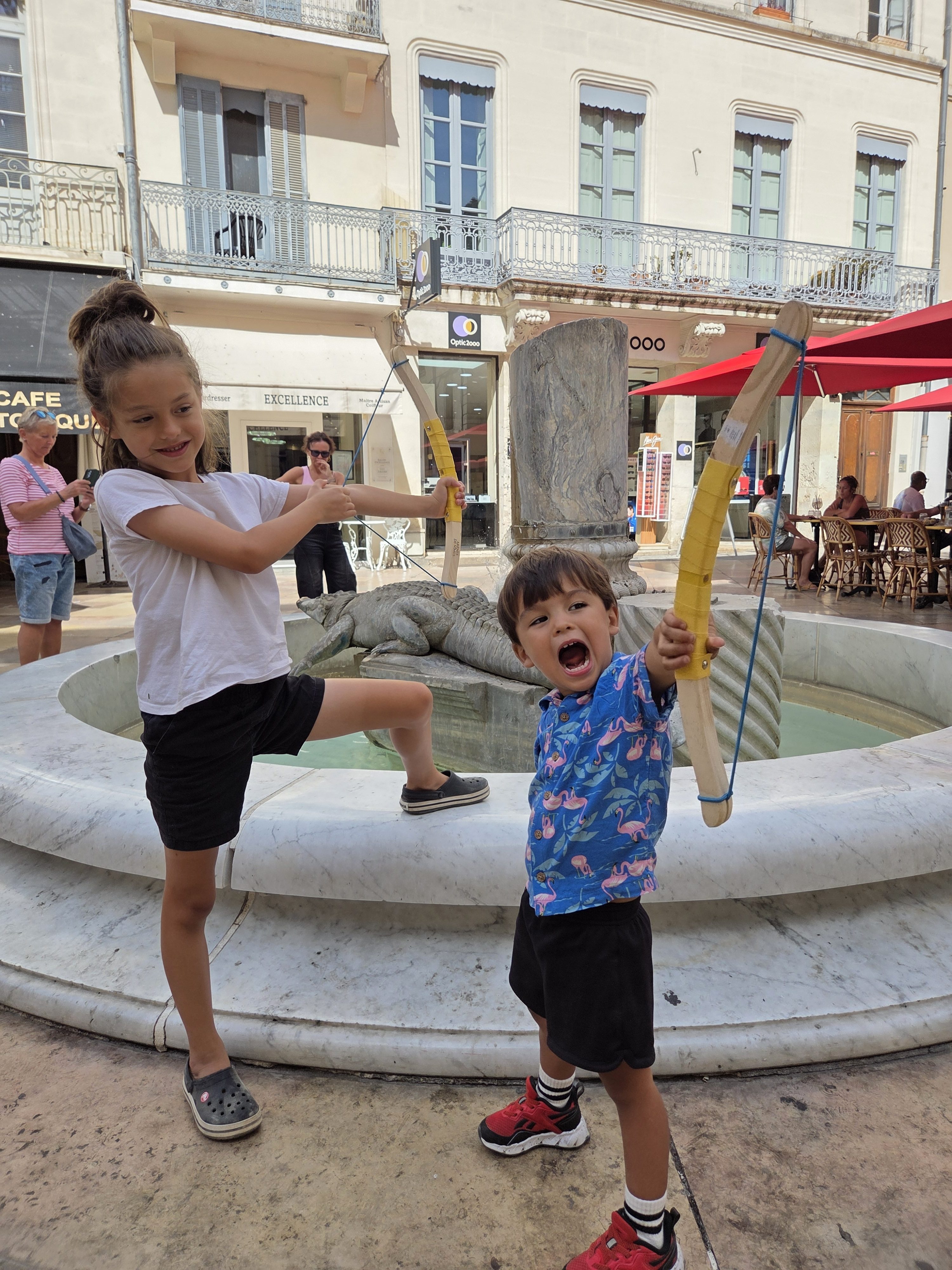 Remi and Arlo posing with their new toy bow-and-arrows in front of a crocodile fountain in a Nîmes square, both grinning and striking warrior poses