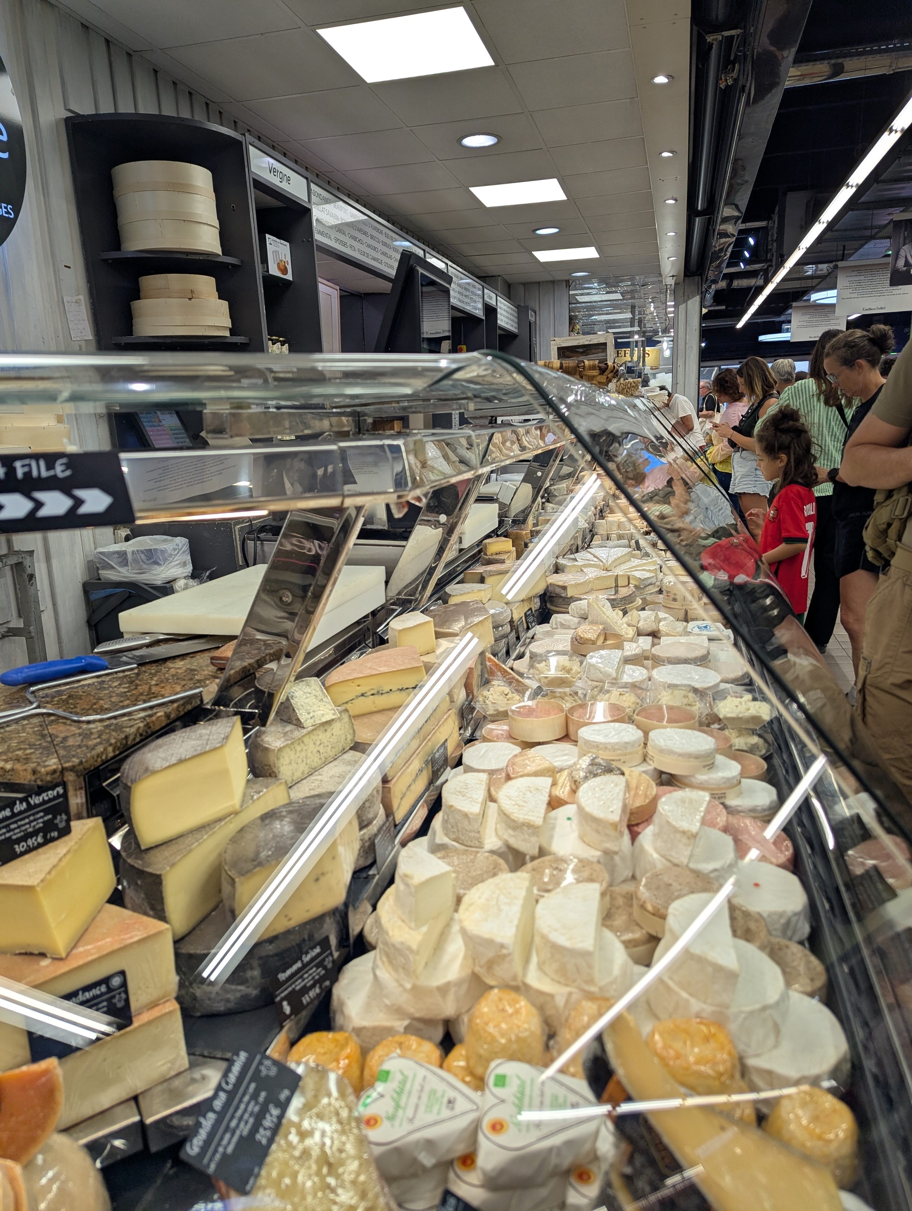 A long glass cheese counter at Les Halles filled with wedges, wheels, and small rounds of cheese, with Remi in a red shirt waiting among other shoppers