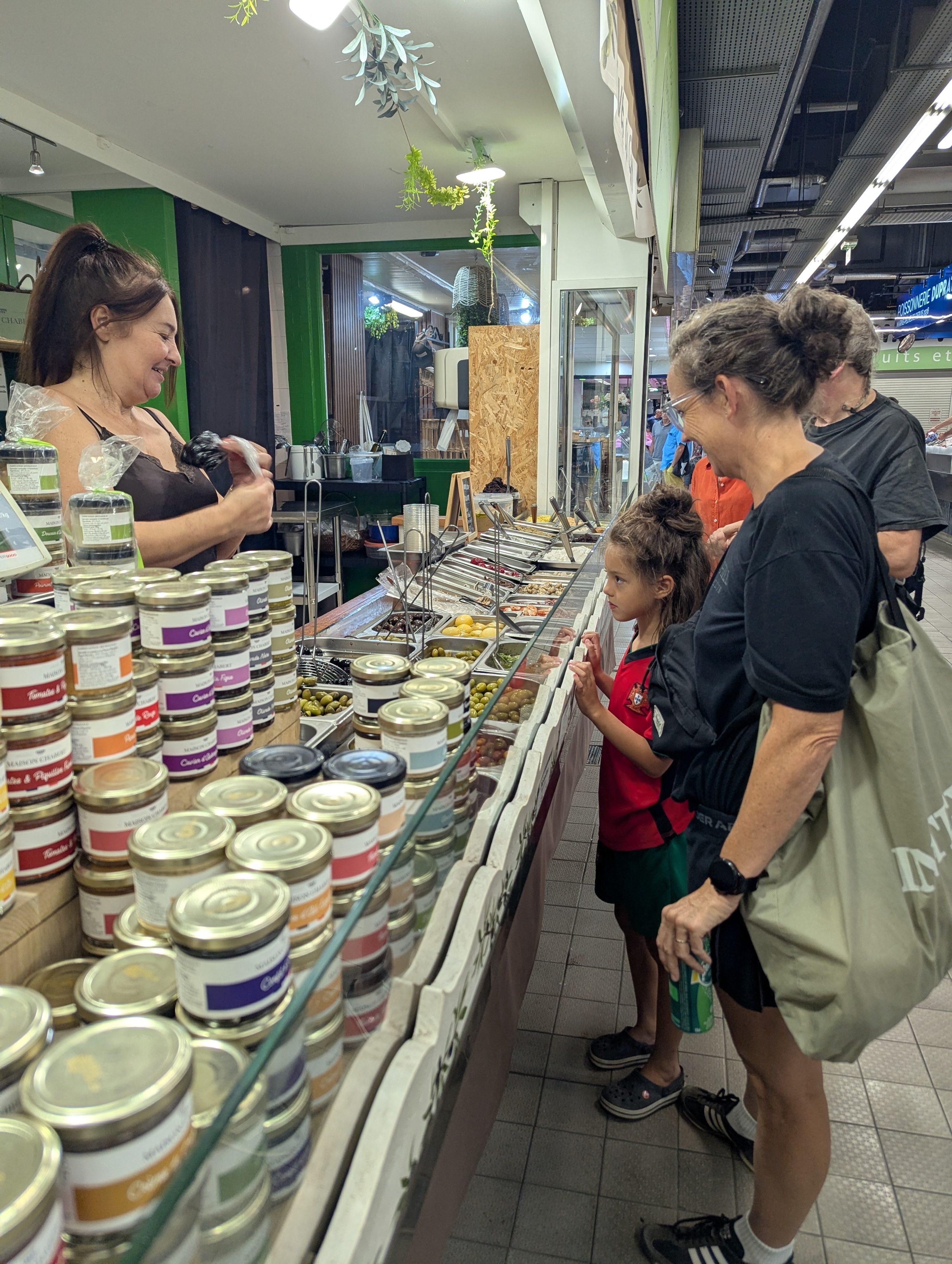 Remi in a red jersey standing beside a parent at an olive and tapenade stall, rows of small jars stacked on the counter and a smiling vendor behind it