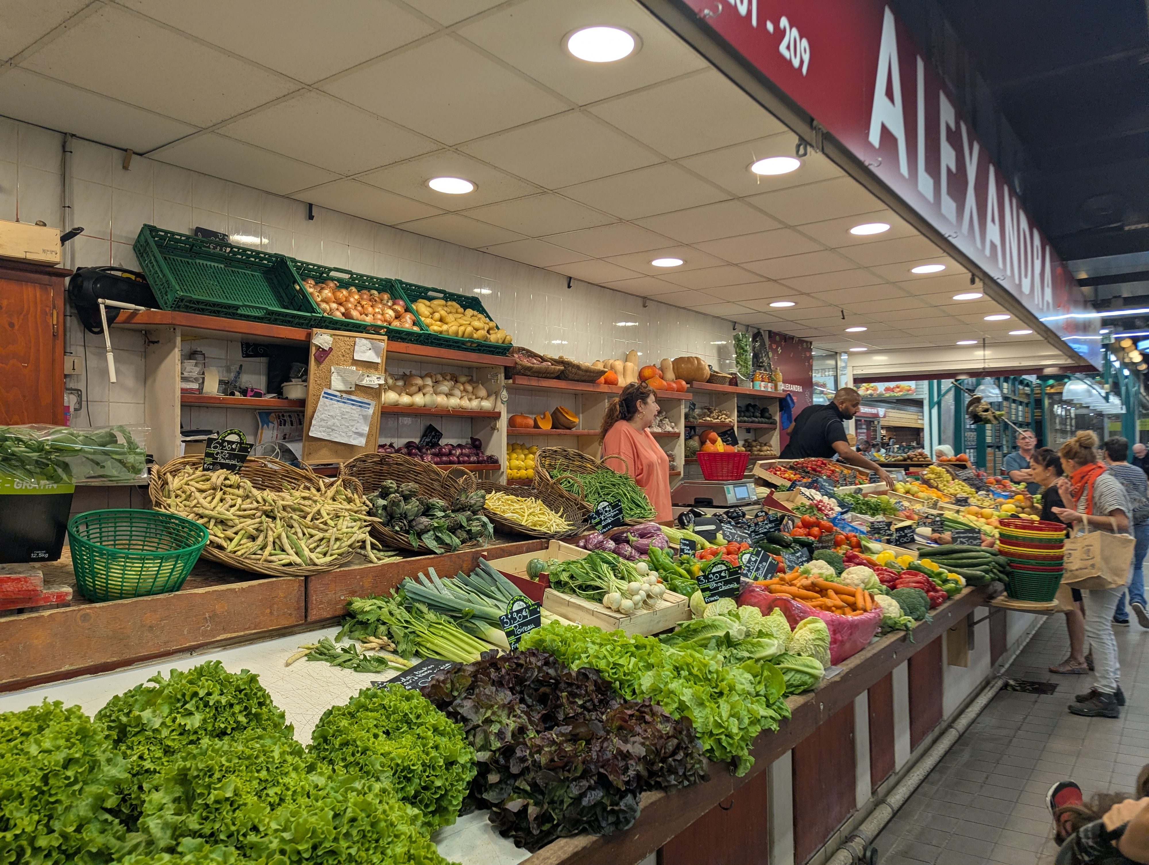A produce stall inside Les Halles de Nîmes with baskets of green beans, lettuces, leeks, and vegetables, and shoppers browsing the aisle beyond