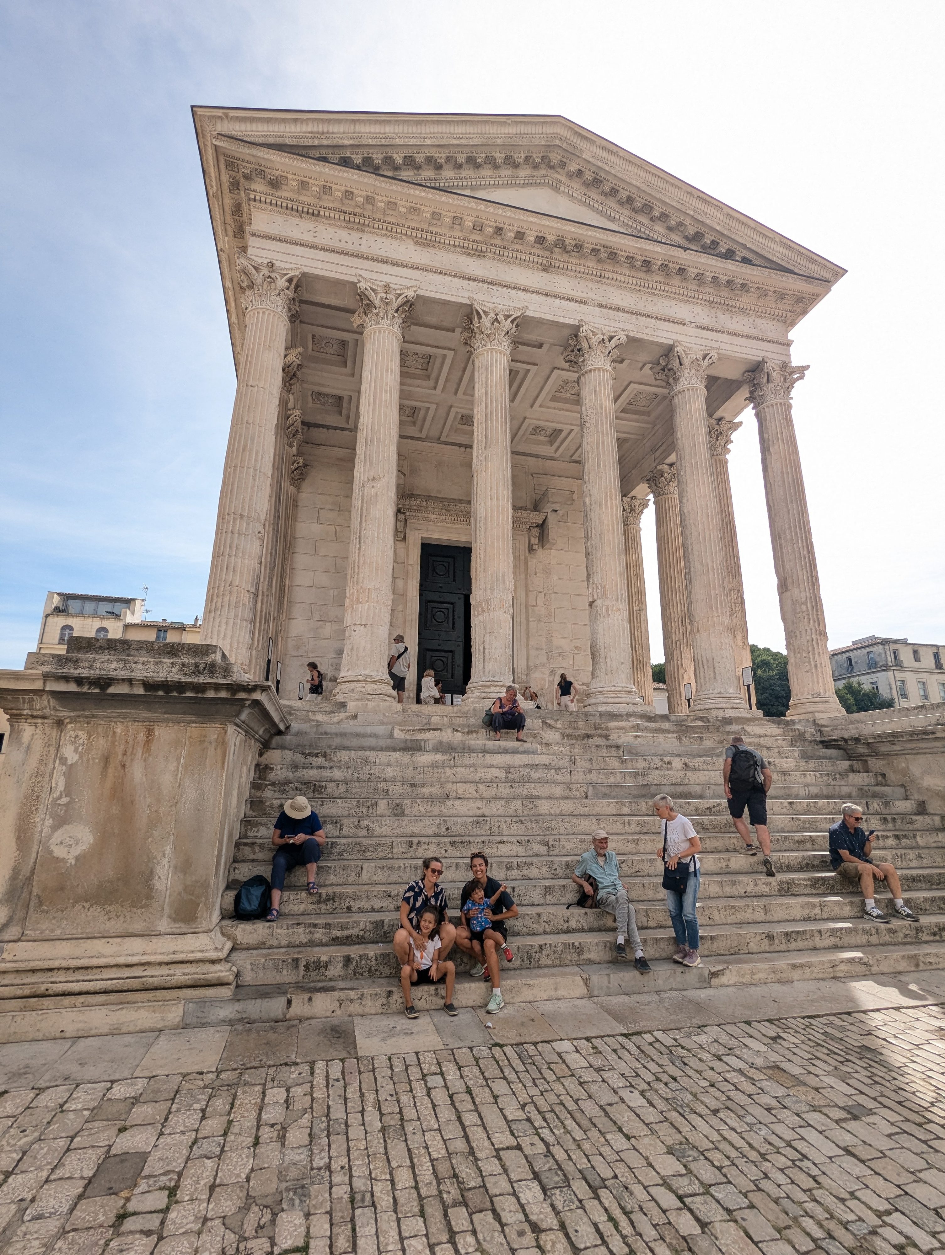 The whole family sitting together on the wide stone steps of the Maison Carrée, the tall Corinthian columns and pediment towering above them