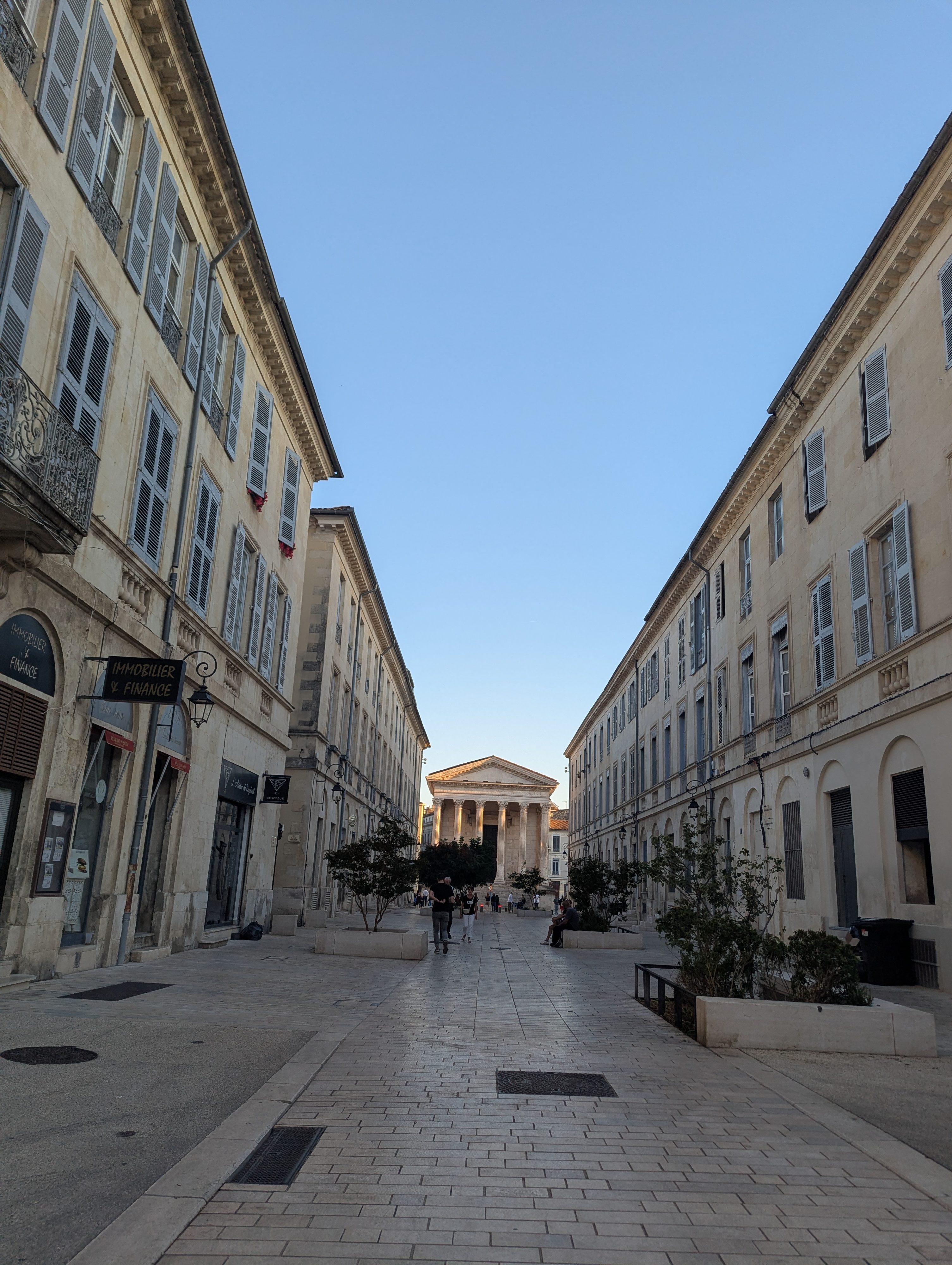 A wide pedestrian boulevard in Nîmes lined with honey-colored stone buildings, the columned facade of the Maison Carrée glowing in the late afternoon sun at the far end