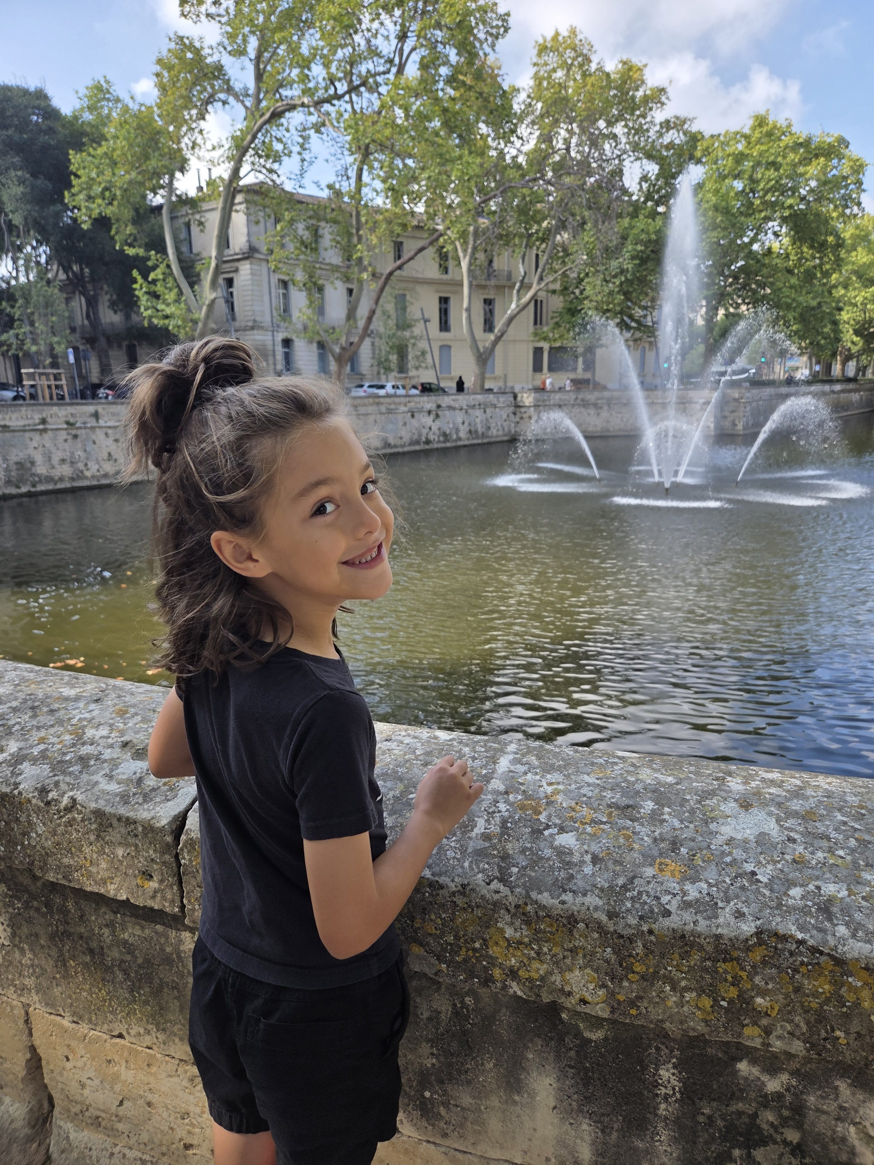 Remi smiling back over her shoulder at the camera as she leans on a mossy stone wall, with fountains spraying in the canal behind her and cream-colored buildings across the water
