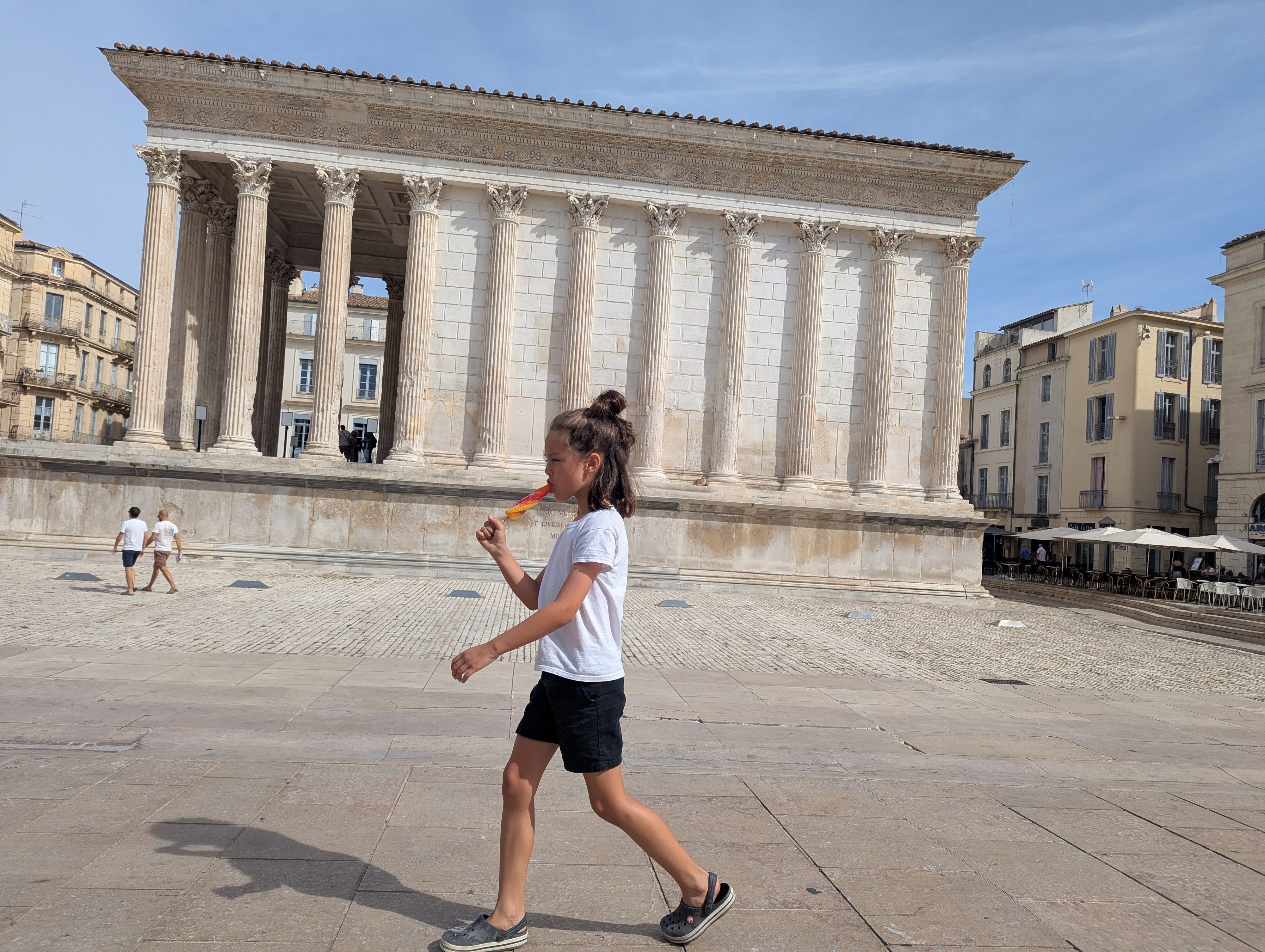 Remi walking past the Maison Carrée eating a rainbow popsicle, the columned facade of the Roman temple filling the background