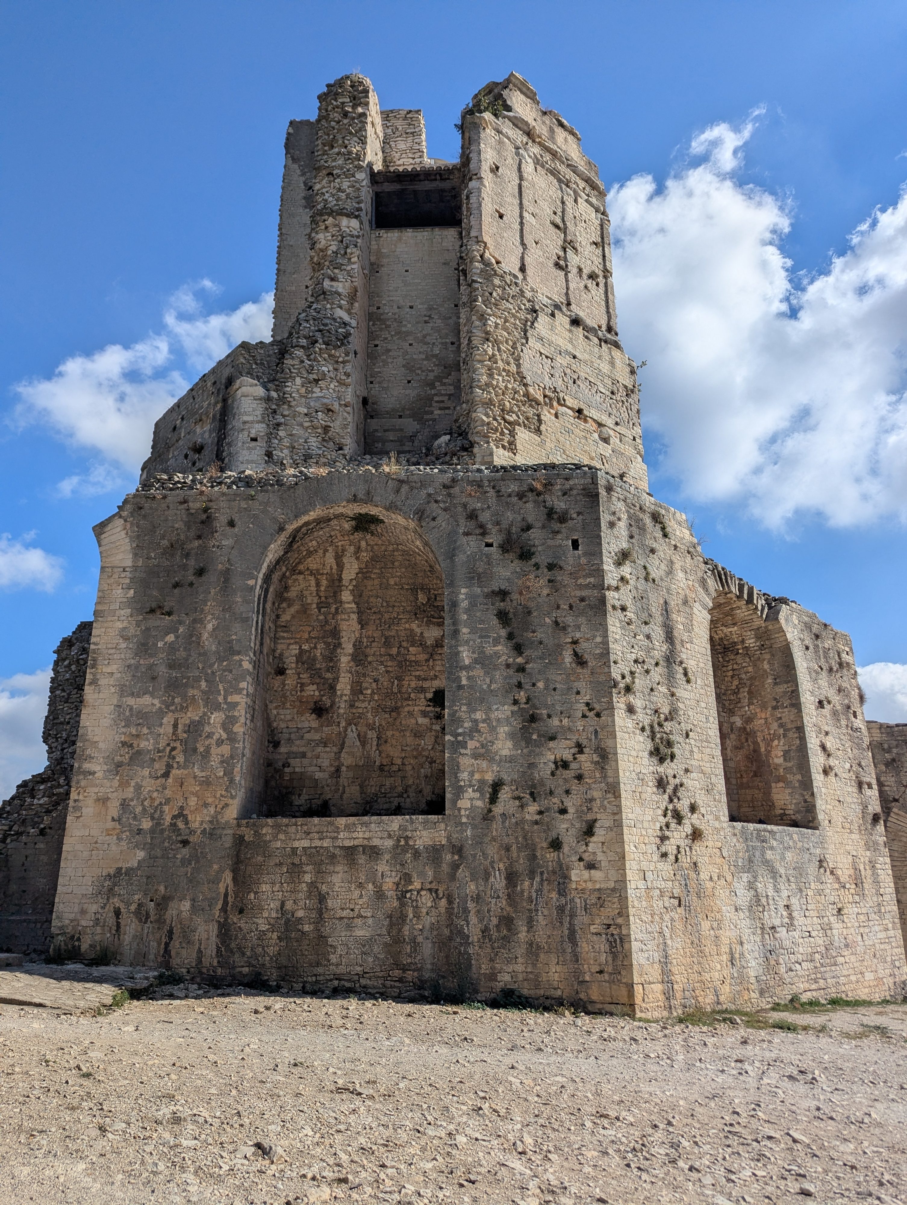The weathered stone walls and arched openings of the Tour Magne rising against a blue sky, seen from the gravel ground at its base